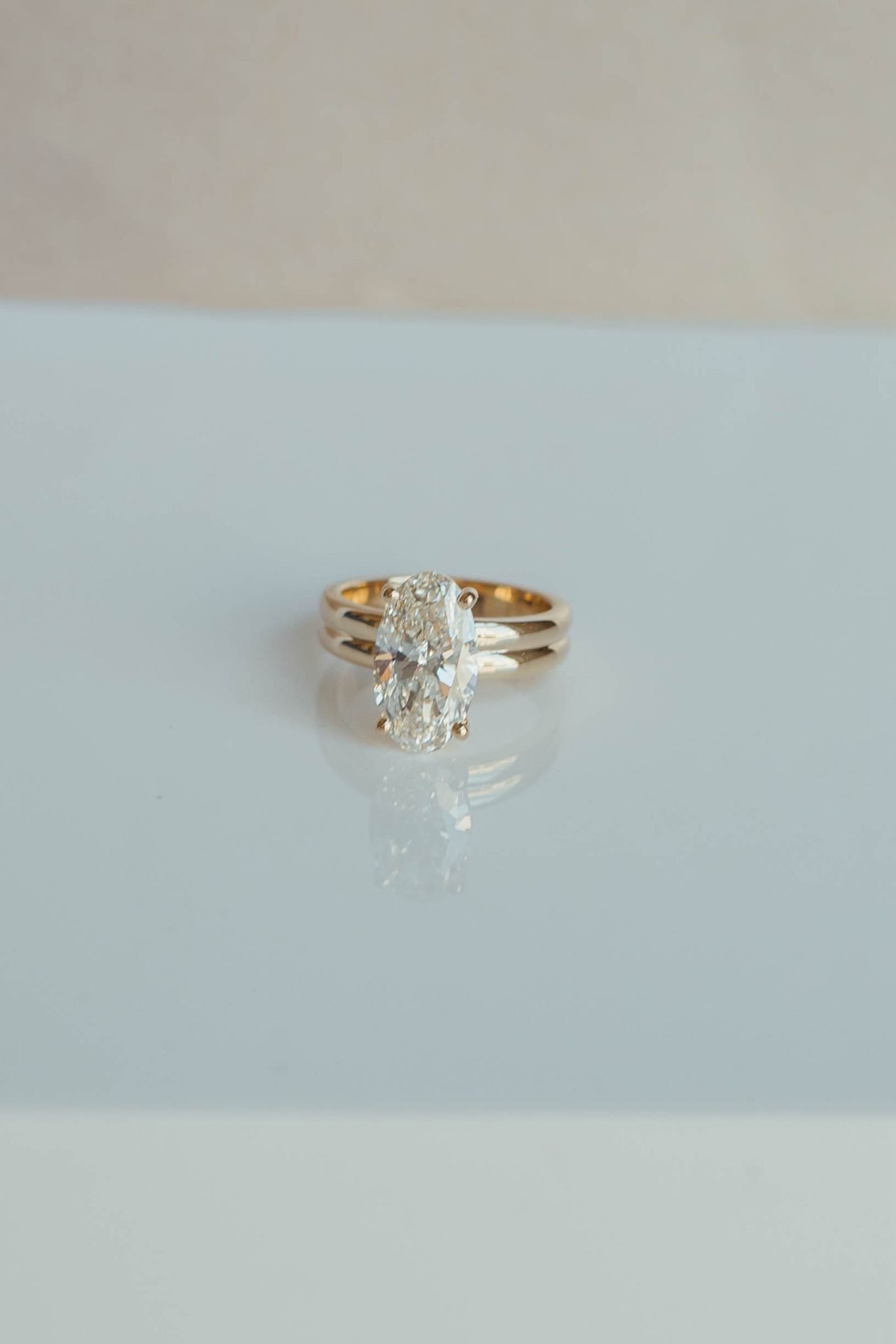 Gold ring with a large oval diamond solitaire on a reflective white surface with a blurred background.