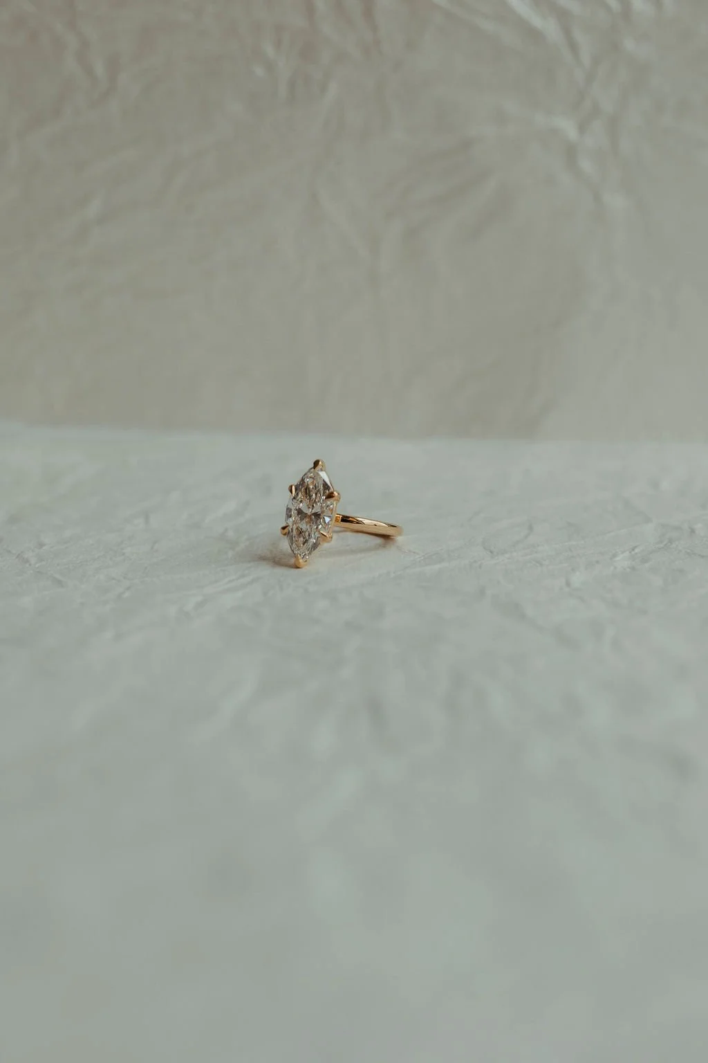 A gold ring with a large marquise-cut diamond solitaire on a textured white surface with a textured wall background.
