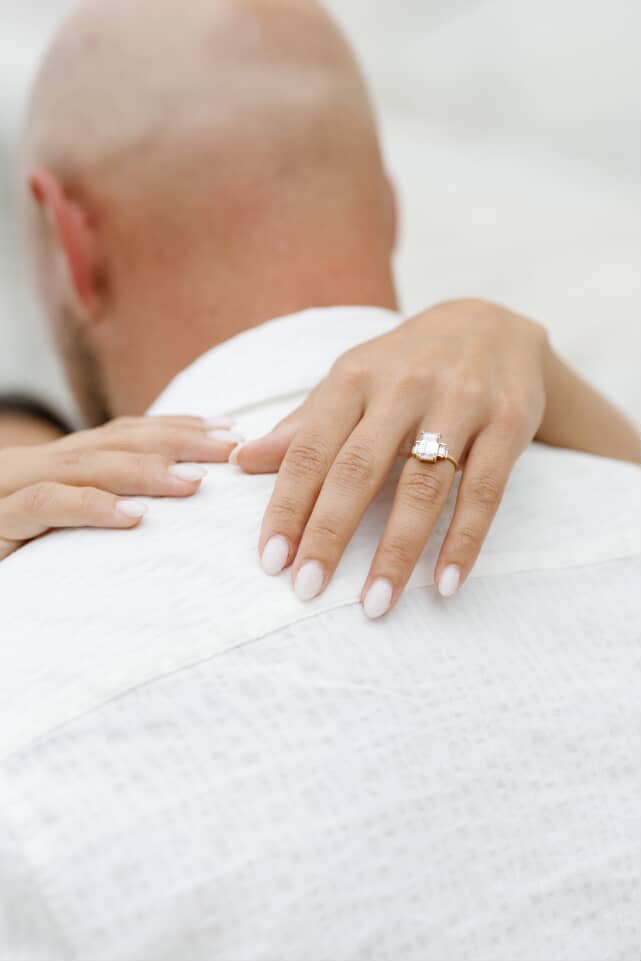 Close-up of a woman's hand with a diamond engagement ring on her ring finger, resting on a man's shoulder who has a shaved head, in a white shirt or gown.