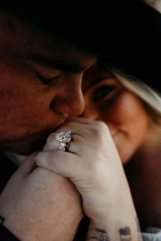 Close-up of a man and woman holding hands. The woman's hand shows a large engagement ring. The man is kissing the woman's hand, and both are wearing dark hats. Blurred background.