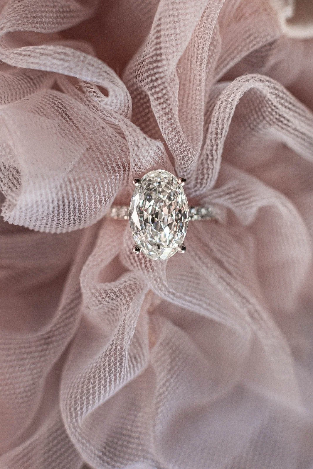 Close-up of an engagement ring with a large oval cut diamond, set on a delicate band, resting on pink mesh fabric.
