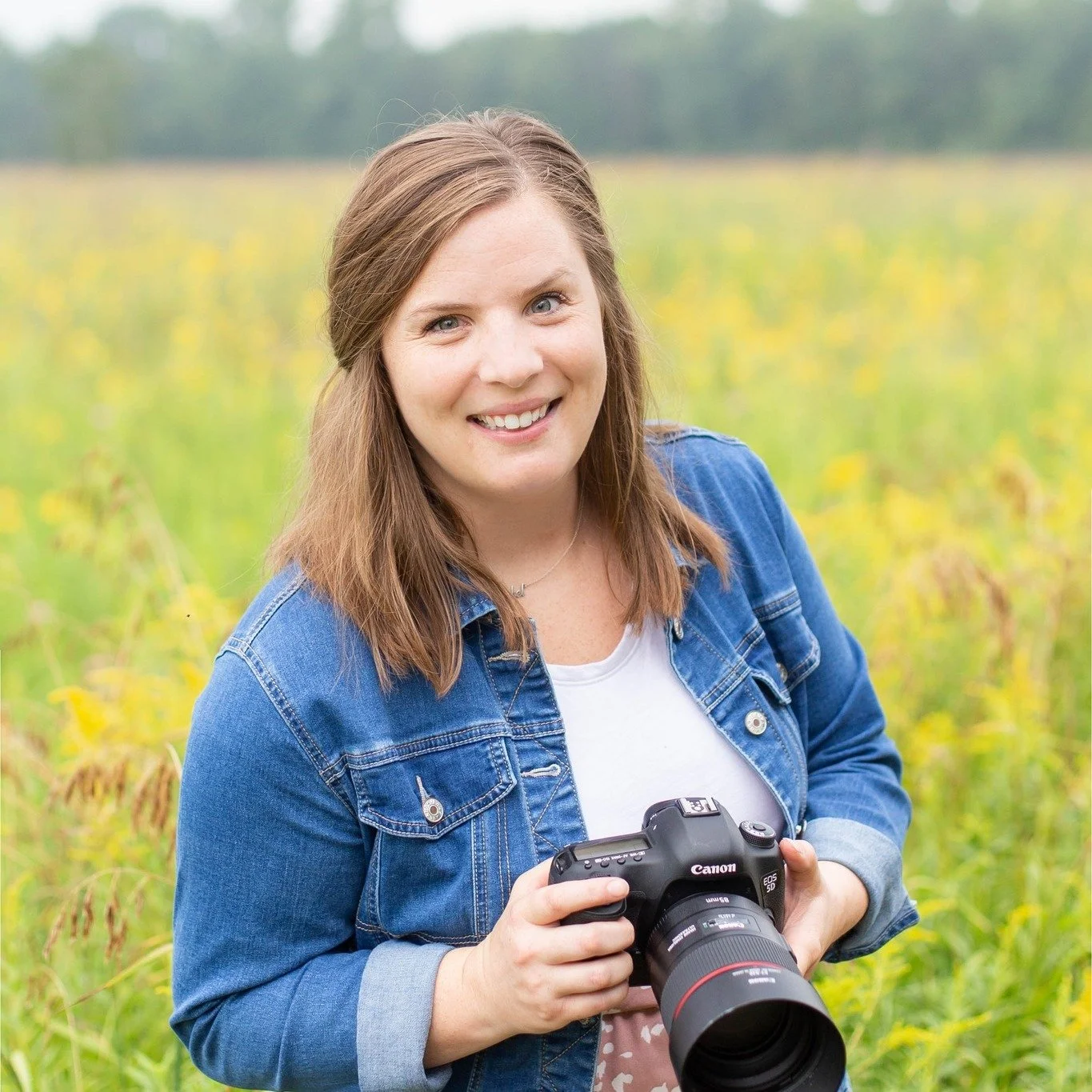 Woman smiling outdoors holding a camera in a field of yellow flowers