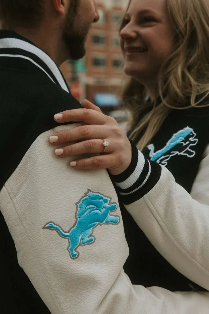 Close-up of a woman and man smiling and embracing, showing a woman wearing a ring on her finger. They are wearing black and white jackets with the Detroit Lions logo, and the background shows city buildings.