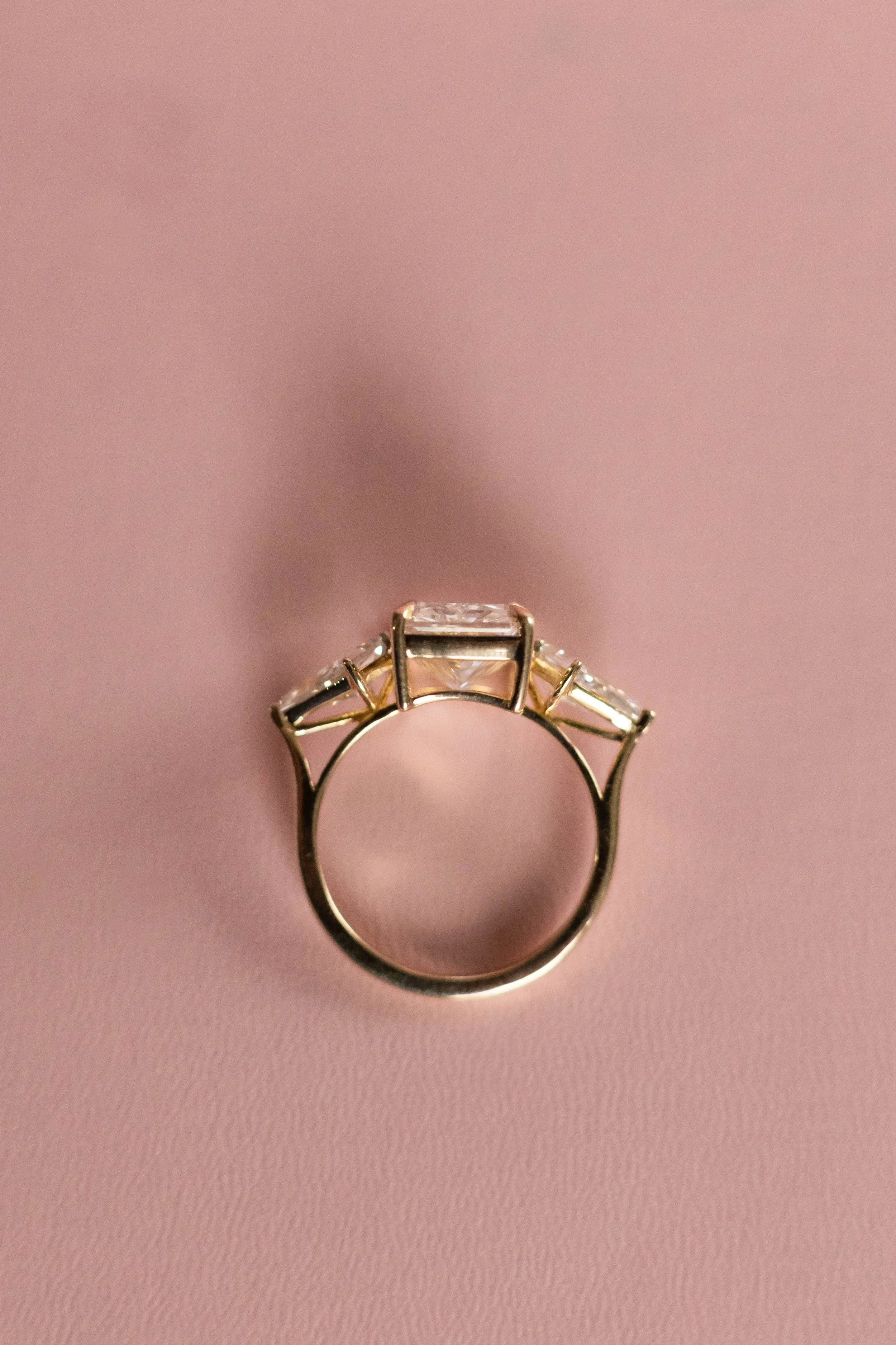Gold ring with rectangular gemstone and smaller side stones on a pink background.