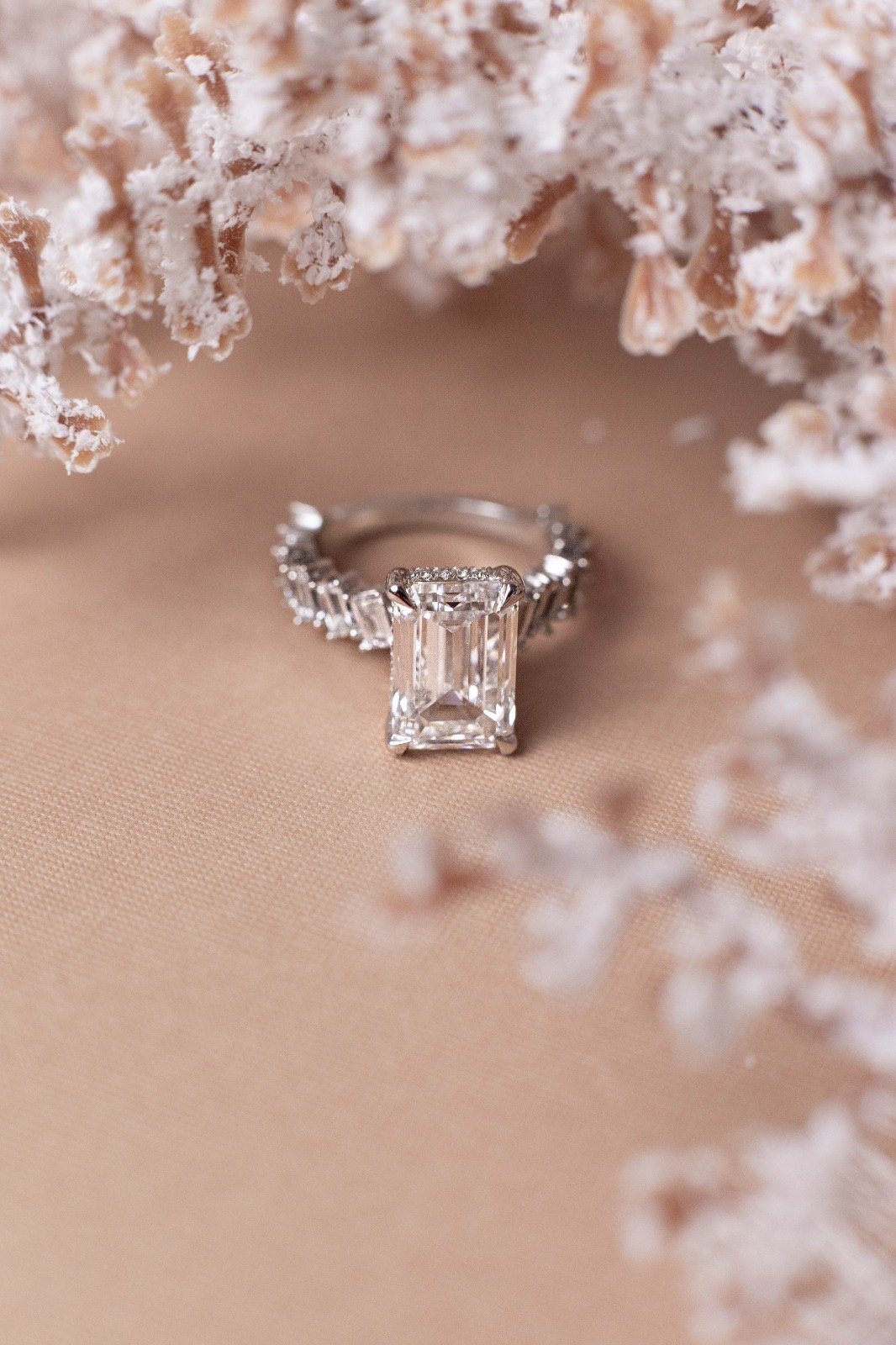 A diamond ring with an emerald-cut center stone and diamond accents on the band, placed on a beige surface surrounded by frosted or snow-covered branches.
