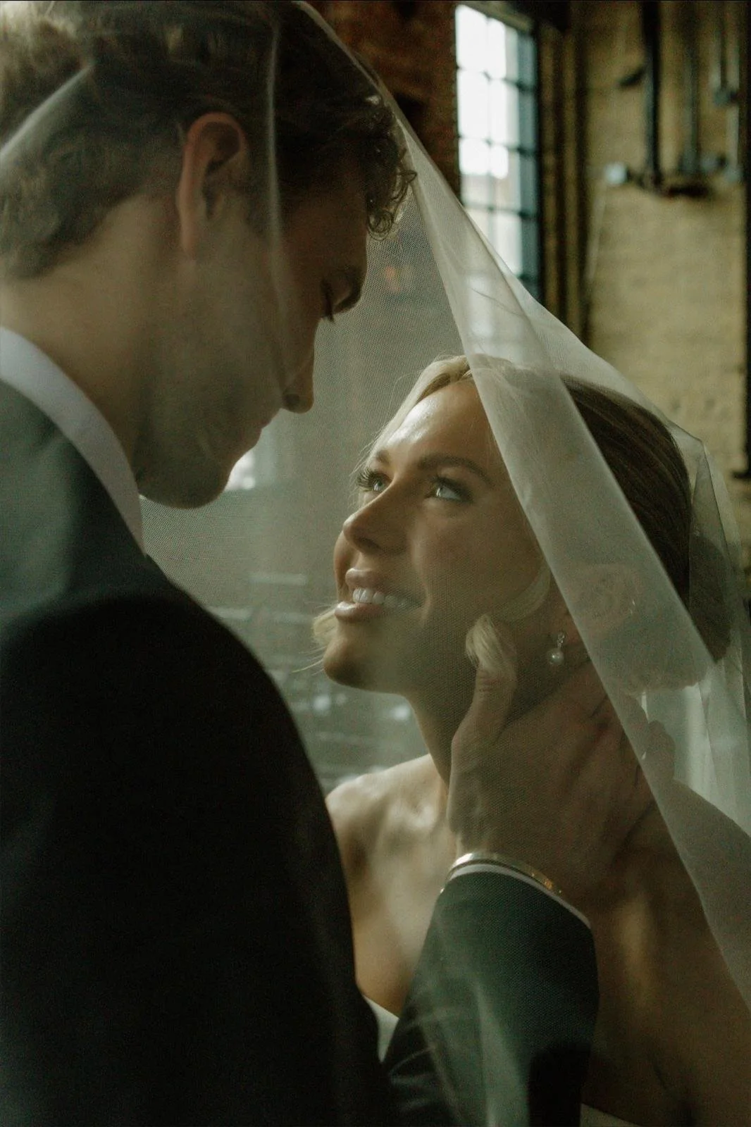 A bride and groom sharing an intimate moment under a veil in a rustic indoor setting with large windows.