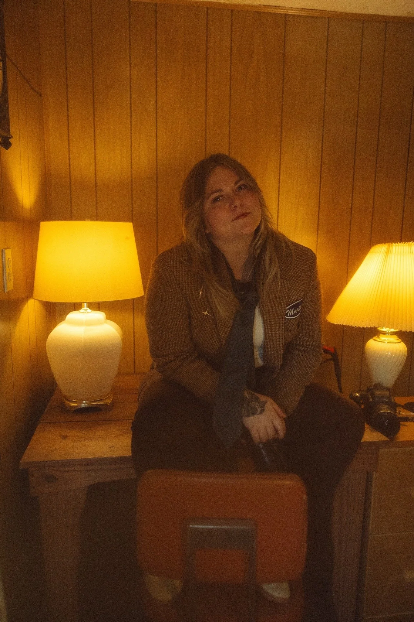 A woman with long, light brown hair sits on a wooden dresser in a cozy, wood-paneled room. She is wearing a brown jacket with a patch that says "Mama," and a black tie. She holds a small black object in her right hand and looks at the camera with a slight smile. Two table lamps with yellow shades are on either side of her, casting warm light. There is a camera on a small table or dresser on her right.