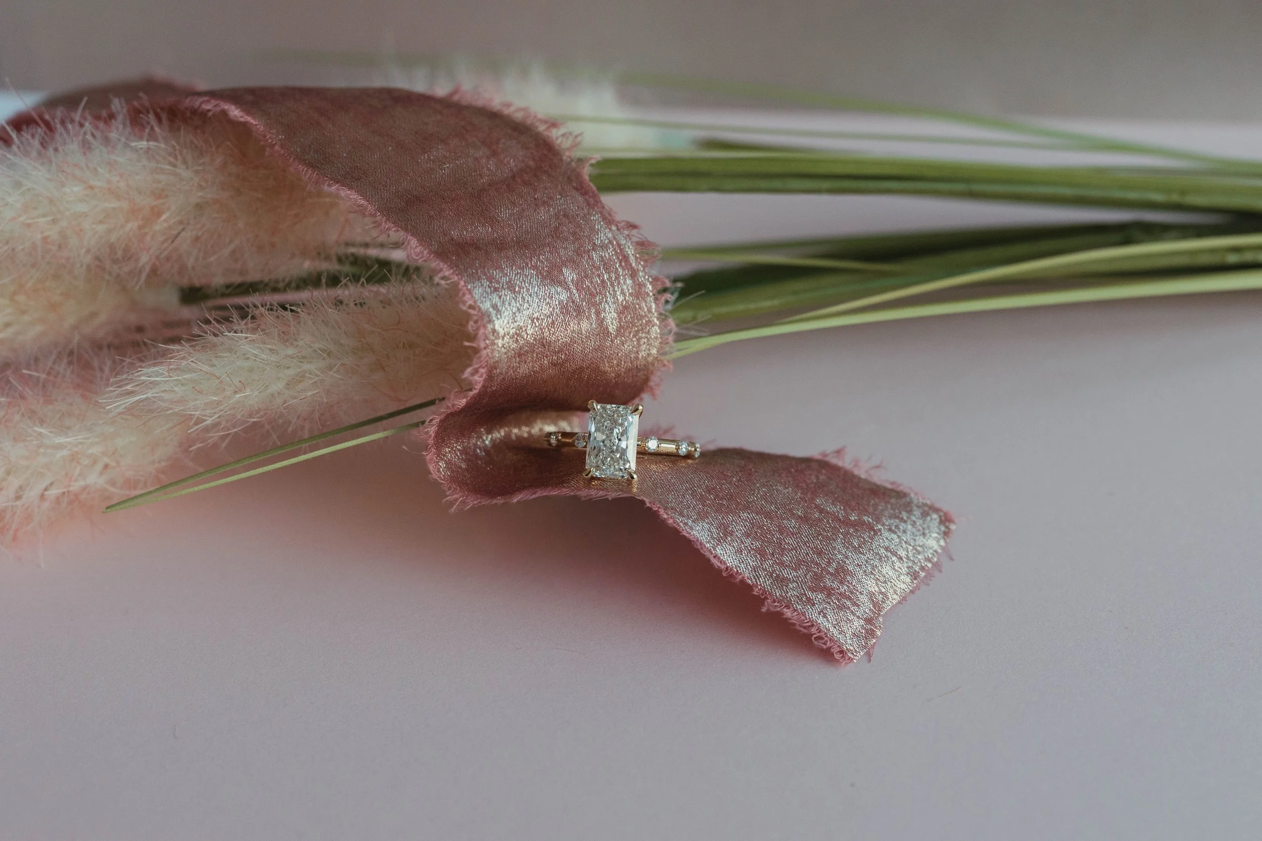 A diamond ring with a rectangular center stone and small side stones resting on a pink, metallic ribbon draped over a light pink surface, with dried pink and white flowers and green grasses as background.