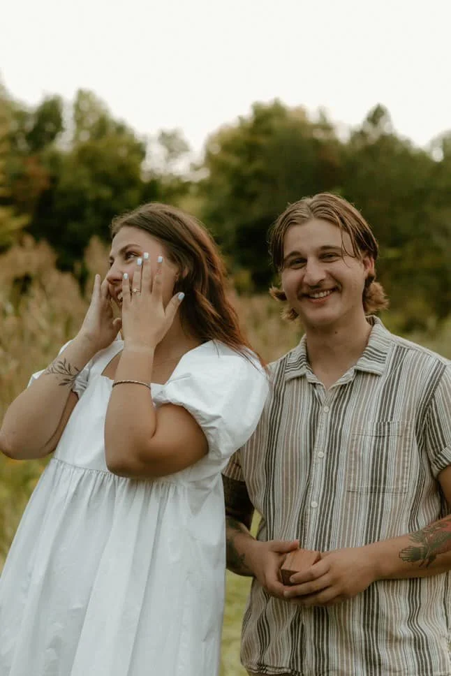 A young woman and man standing outdoors in a natural setting, smiling and enjoying each other's company. The woman is wearing a white dress and holding her face with her hands, while the man is wearing a striped shirt with rolled-up sleeves.