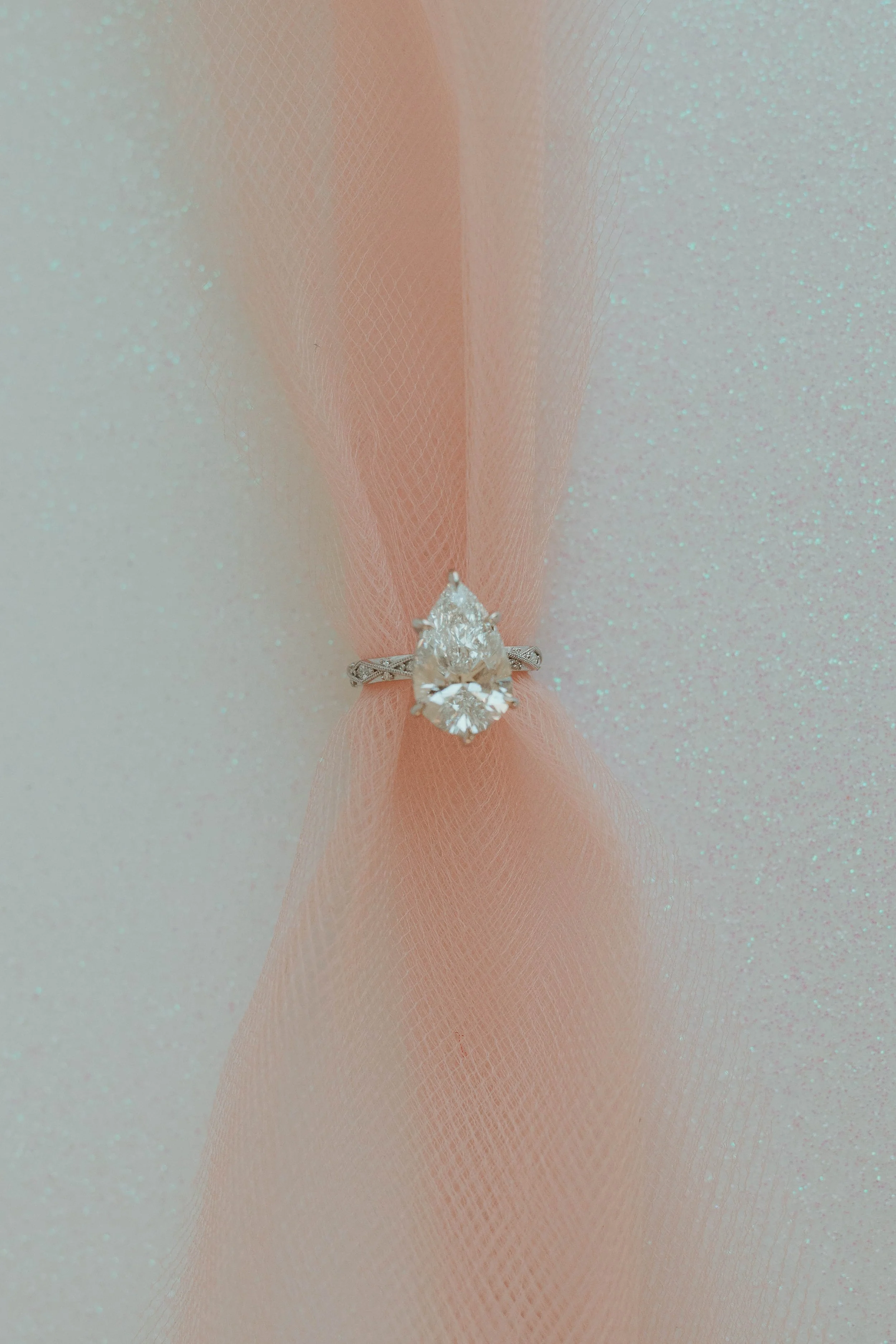 A close-up of a diamond engagement ring displayed against a soft, textured, pastel background with a pinkish hue.