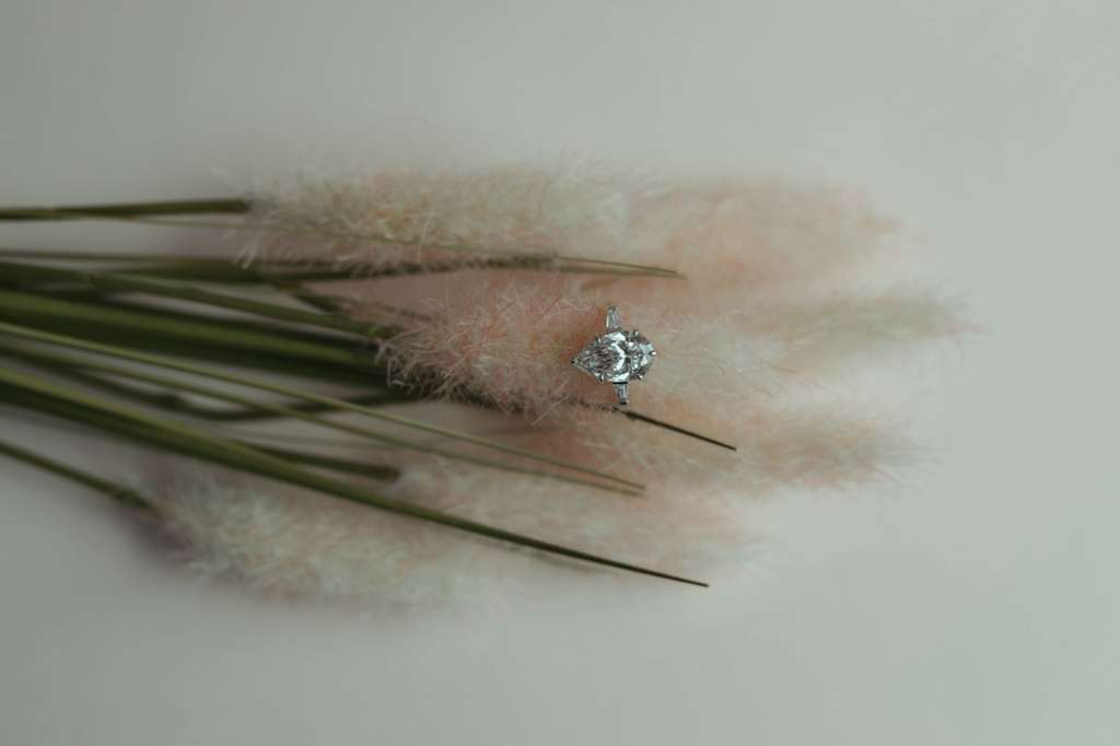 A sparkling diamond engagement ring with a large center stone on a delicate band, resting on soft pink and white fluffy grasses against a plain background.