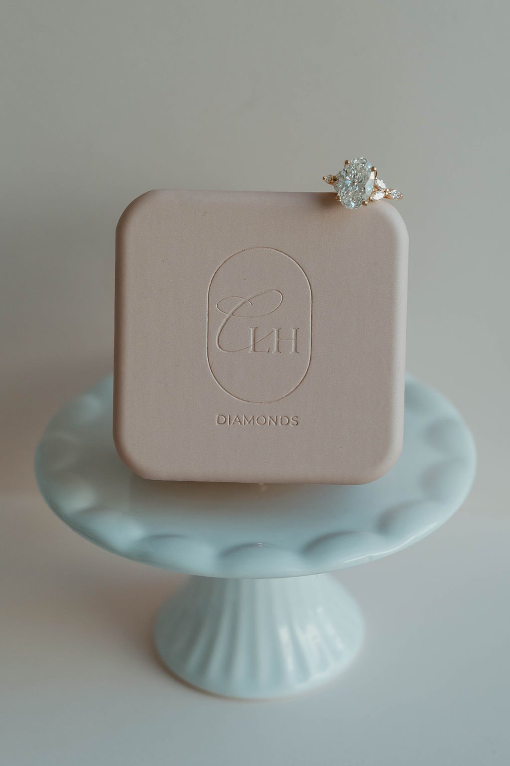 A diamond ring with a large oval diamond and smaller side stones resting on a beige jewelry box that has a logo with the initials 'T H' and the word 'Diamonds.' The jewelry box is placed on a light blue ceramic stand or cake with a scalloped edge.