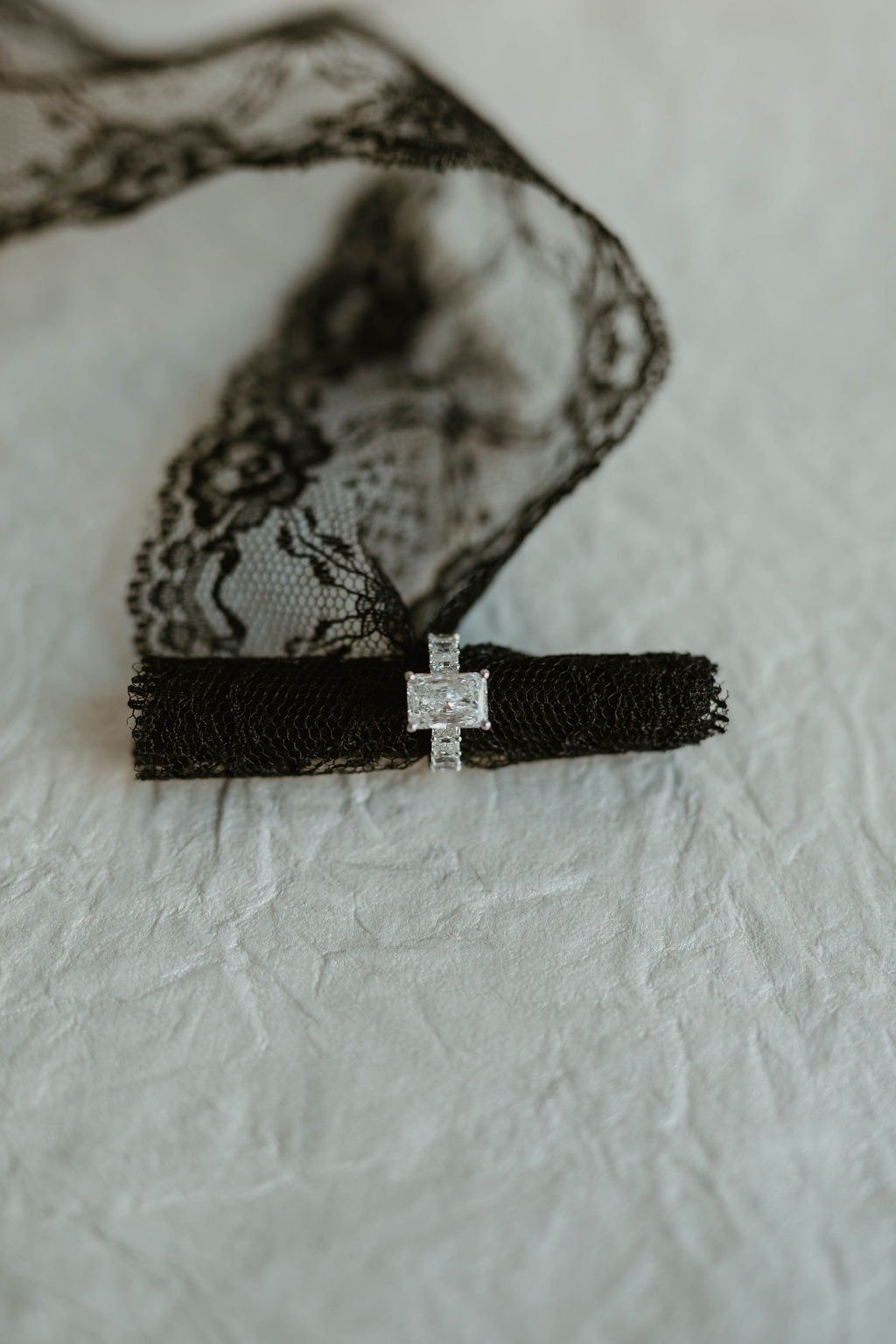 A black lace choker with a square diamond ring in the center, placed on a textured white surface with a black lace fabric in the background.