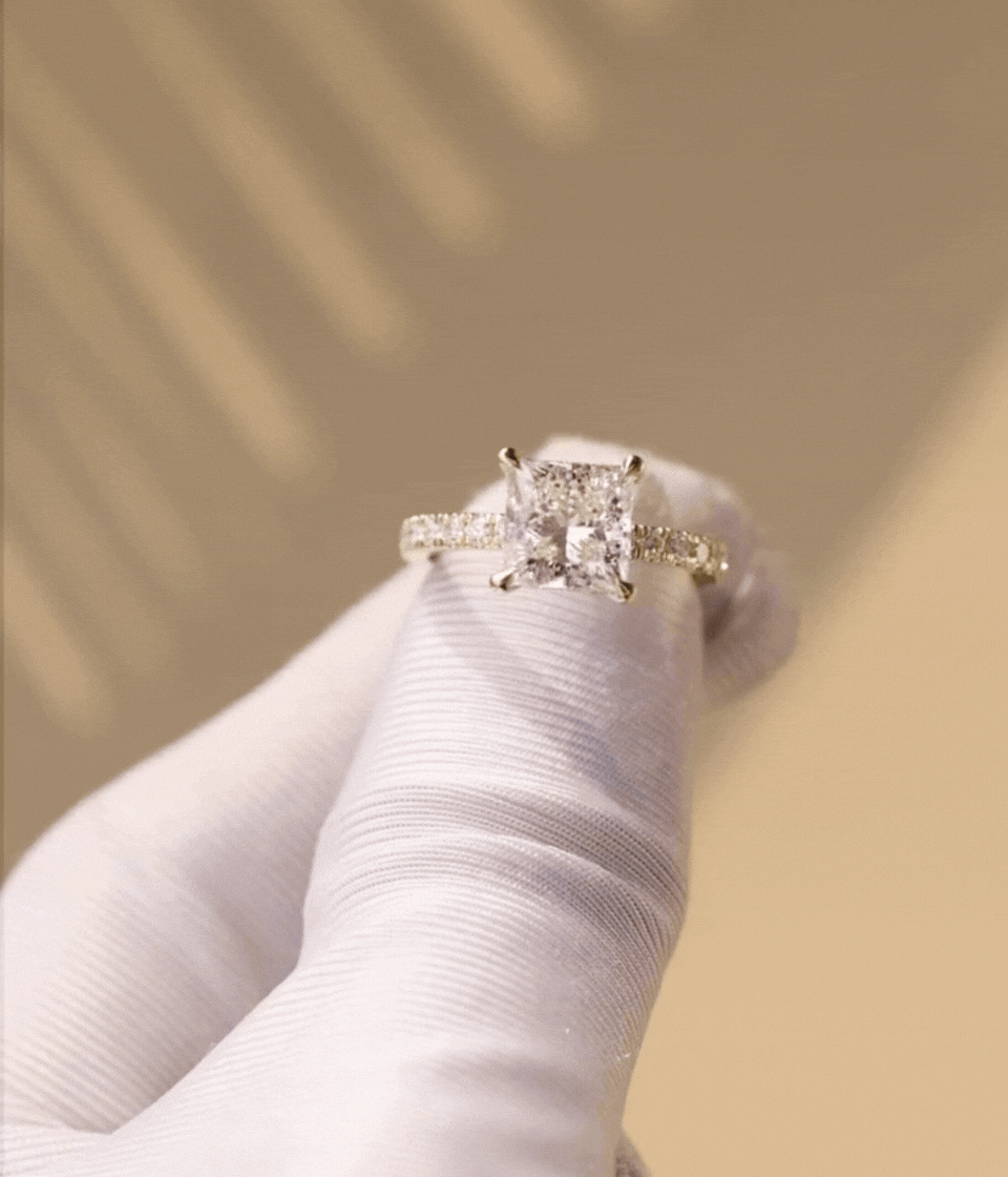 A close-up of a sparkling princess-cut diamond engagement ring with a diamond-studded band, being held by a person wearing a white glove.
