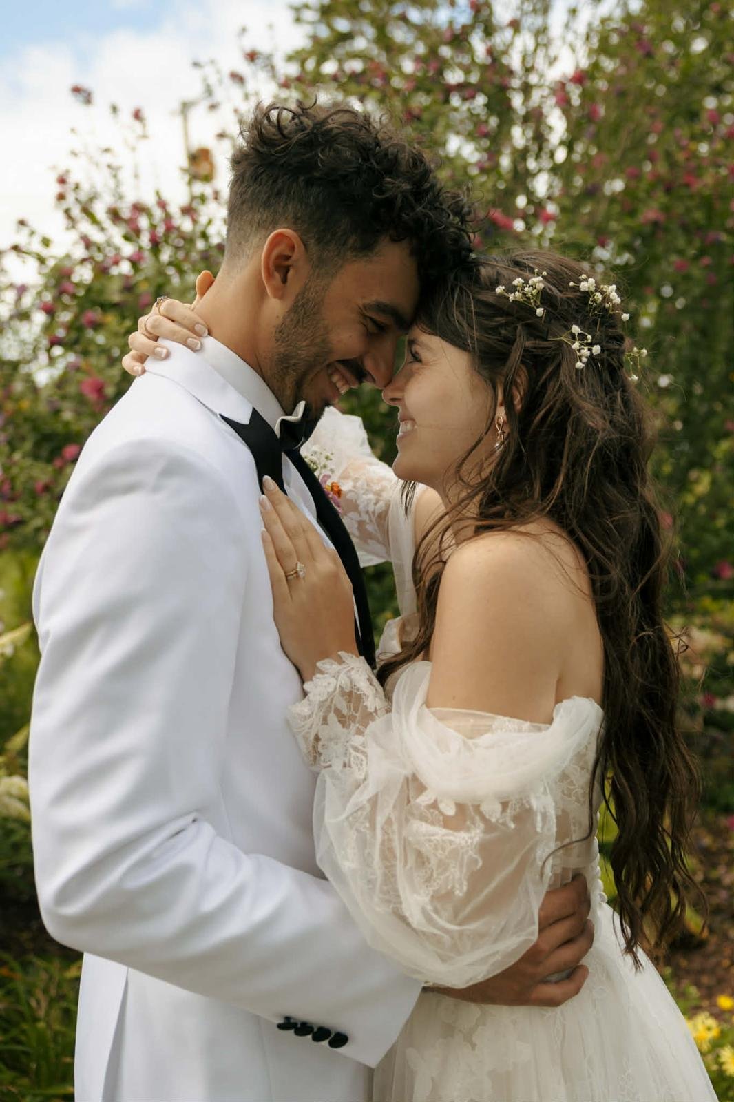 A bride and groom are smiling and touching foreheads in a romantic moment outdoors surrounded by greenery and pink flowers.
