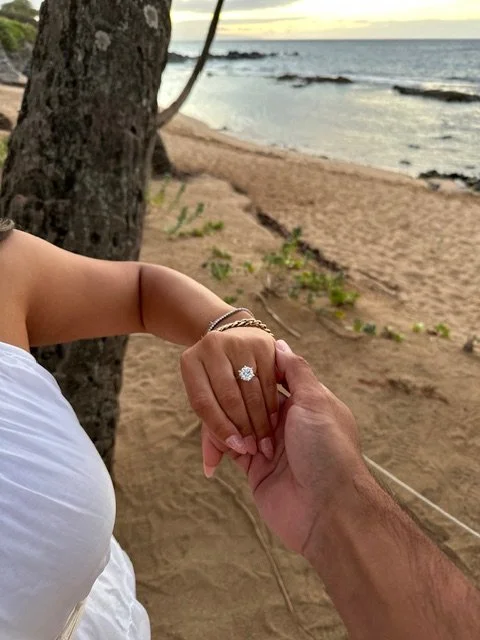 A person with a ring on their finger holding hands with another person on the beach, with trees, sand, and ocean in the background.