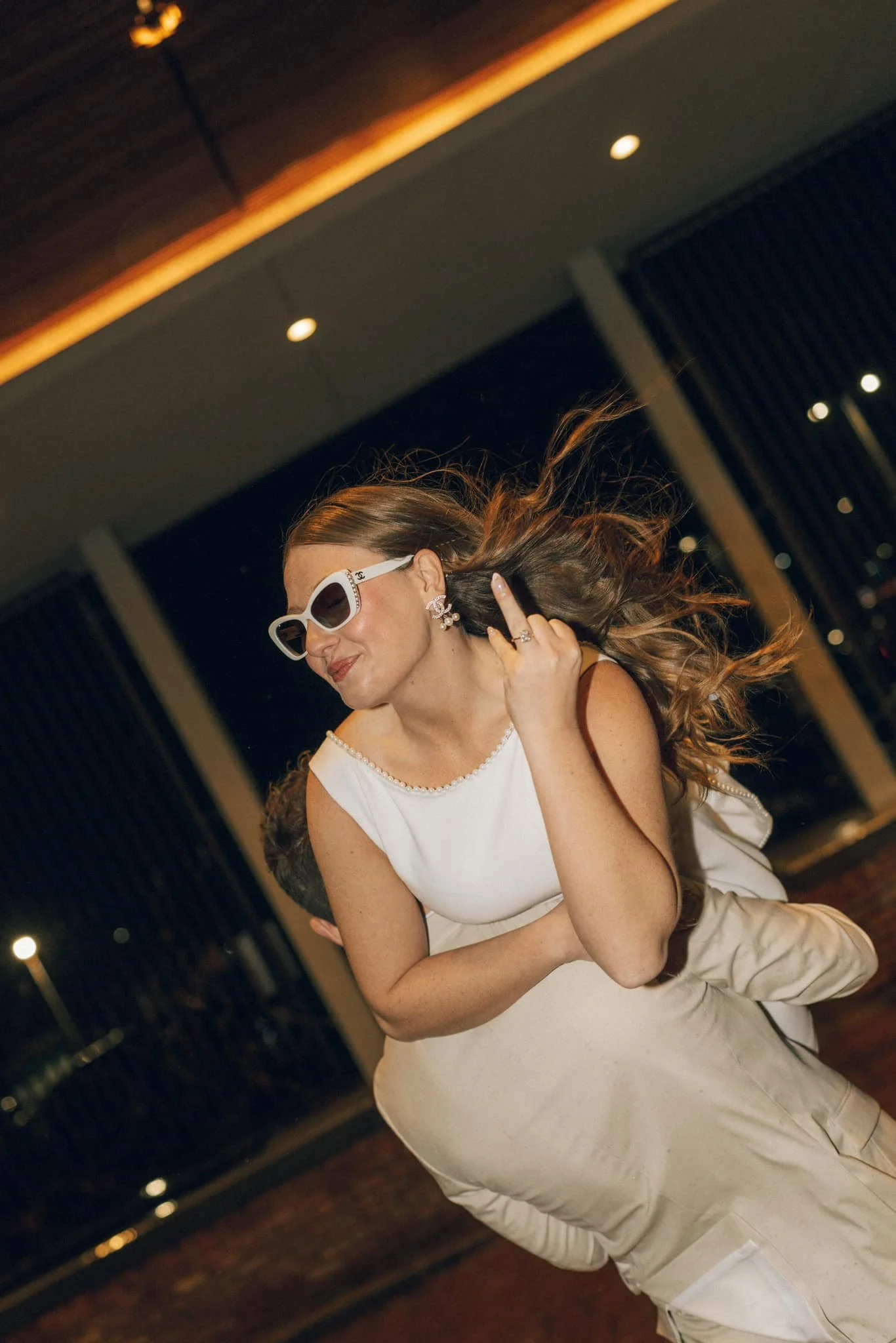 A woman with long hair, wearing sunglasses, earrings, and a white dress, is dancing and having fun at an indoor event during nighttime.