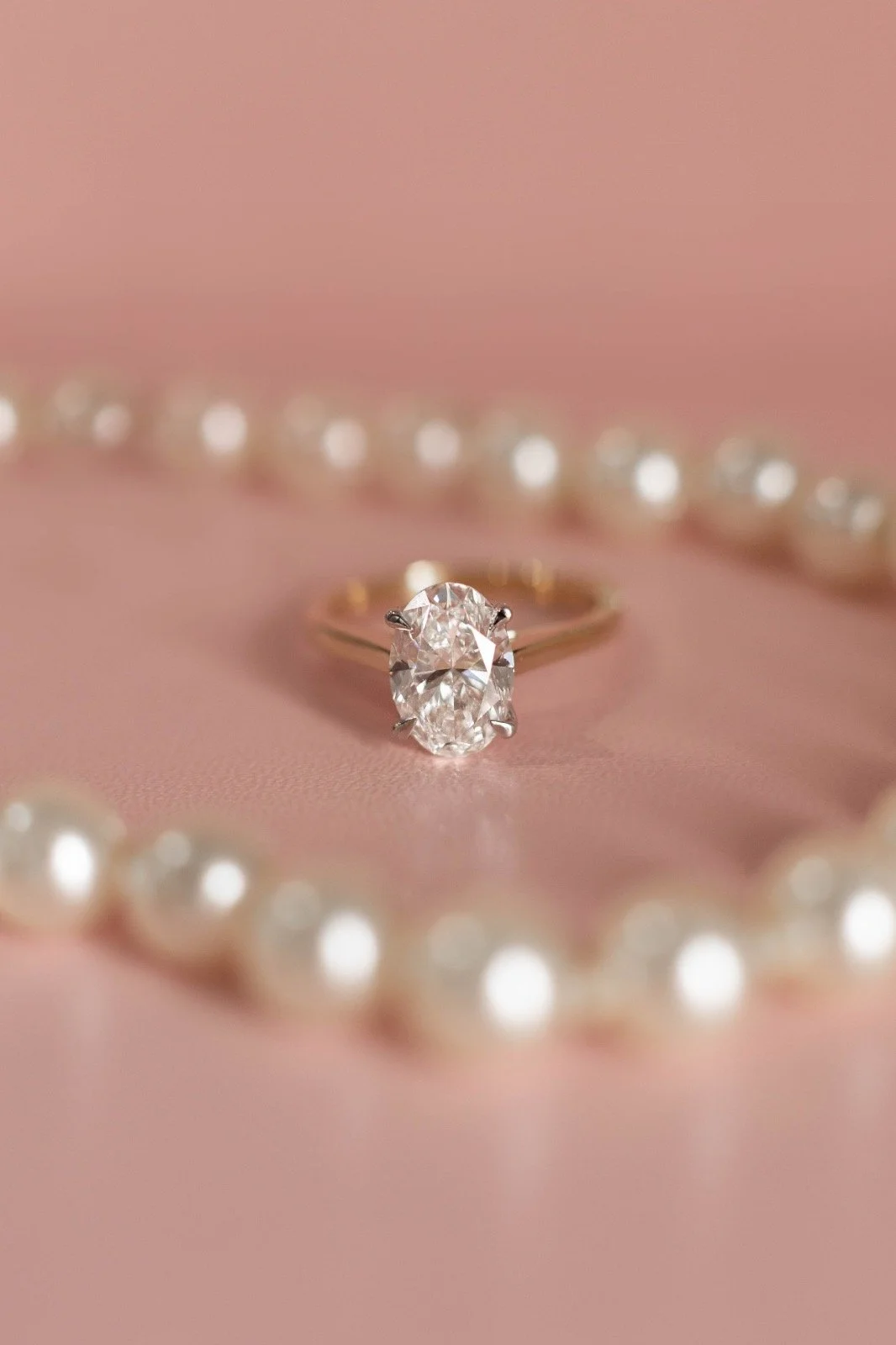 Close-up of an oval diamond engagement ring with a gold band, surrounded by a pearl necklace on a pink background.