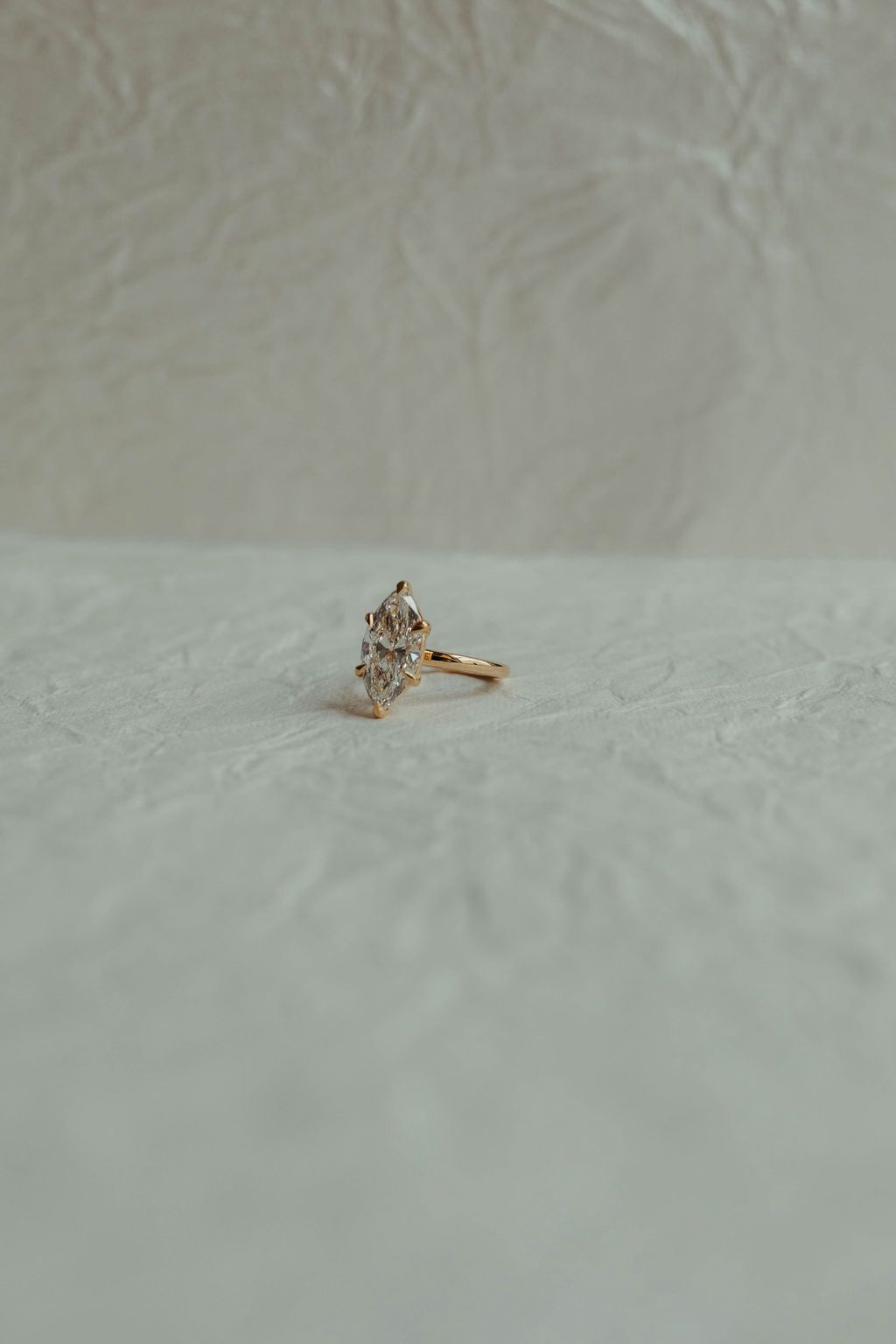 A diamond ring with a marquise-shaped diamond set in a gold band placed on a textured, ivory surface with a plain, textured backdrop.