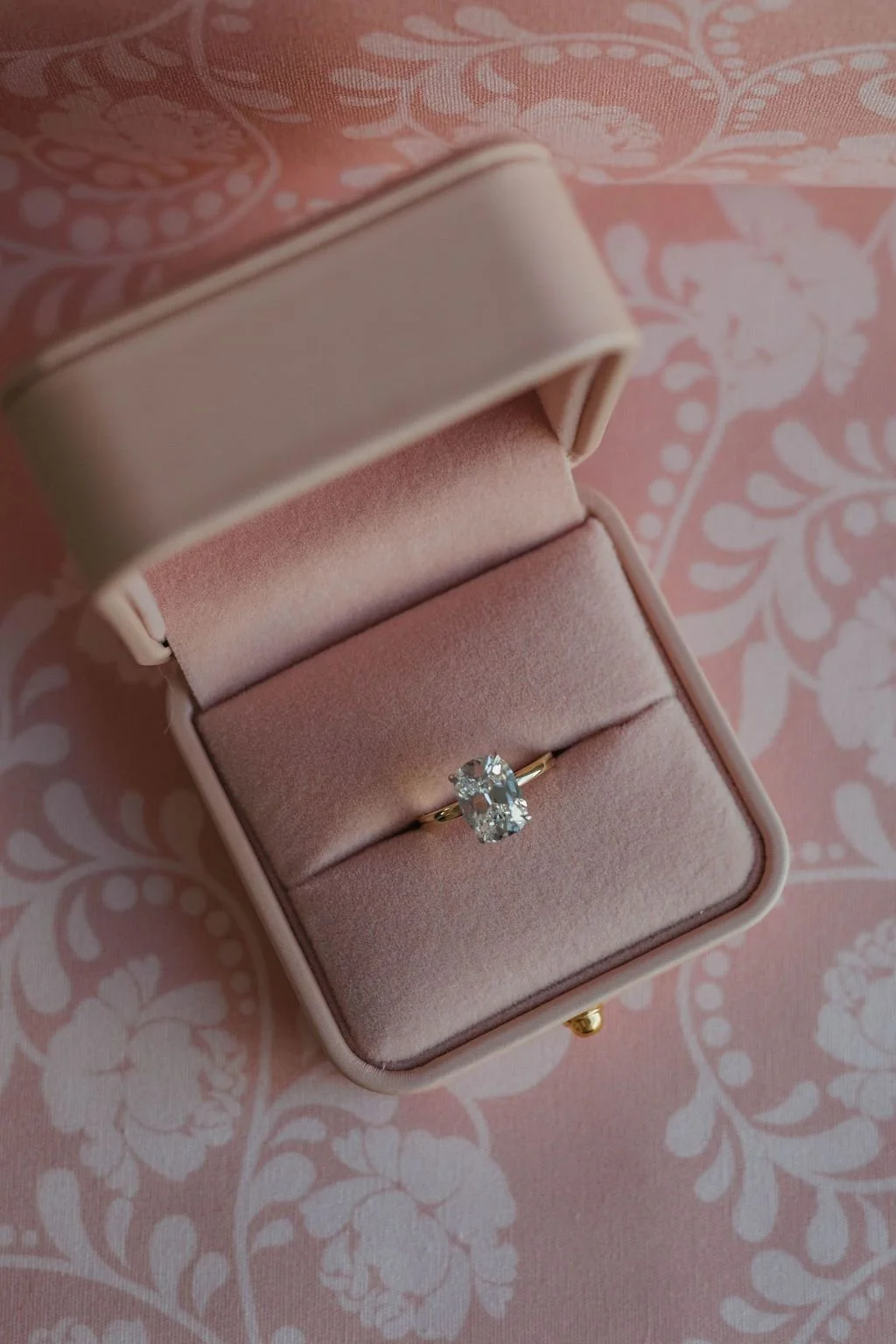 A diamond engagement ring with an oval-shaped center stone in a pink velvet jewelry box, on pink patterned fabric.