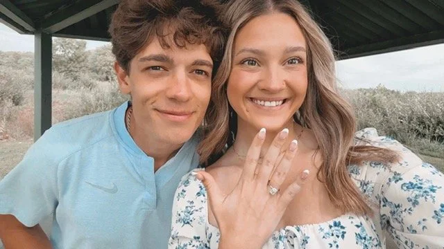 A young couple smiling outdoors, the woman showing off her engagement ring on her finger.