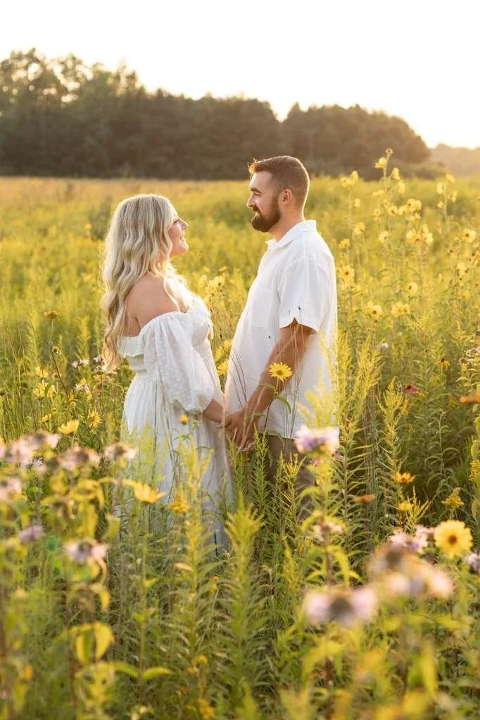 A couple holding hands and smiling at each other in a field of yellow wildflowers during sunset.