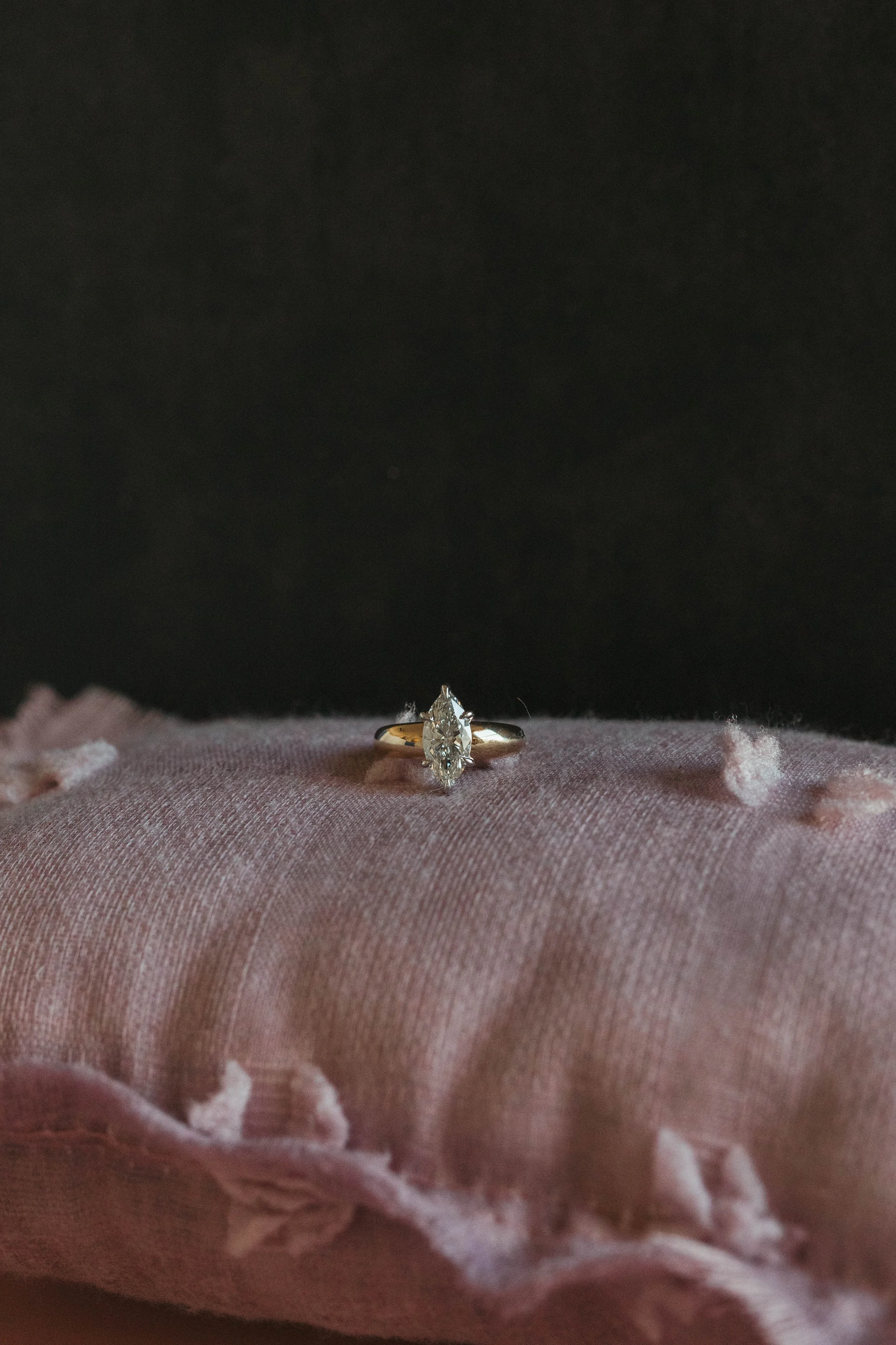 A diamond engagement ring resting on a pink, ruffled fabric cushion against a dark background.