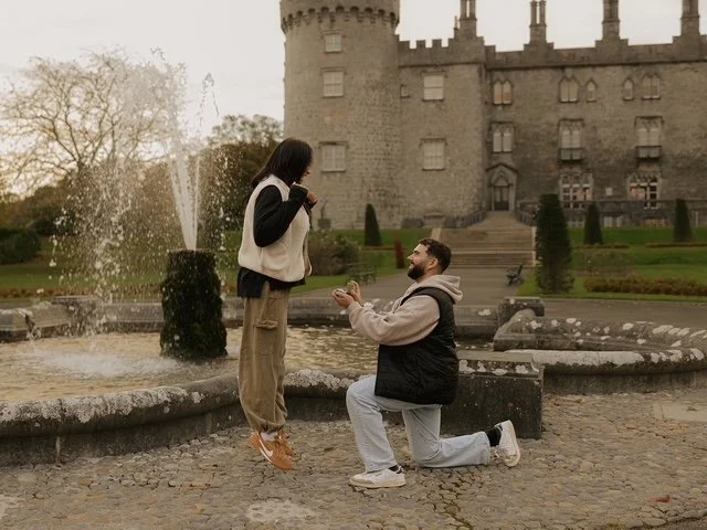 A man is kneeling and proposing to a woman in front of a castle with a fountain nearby.