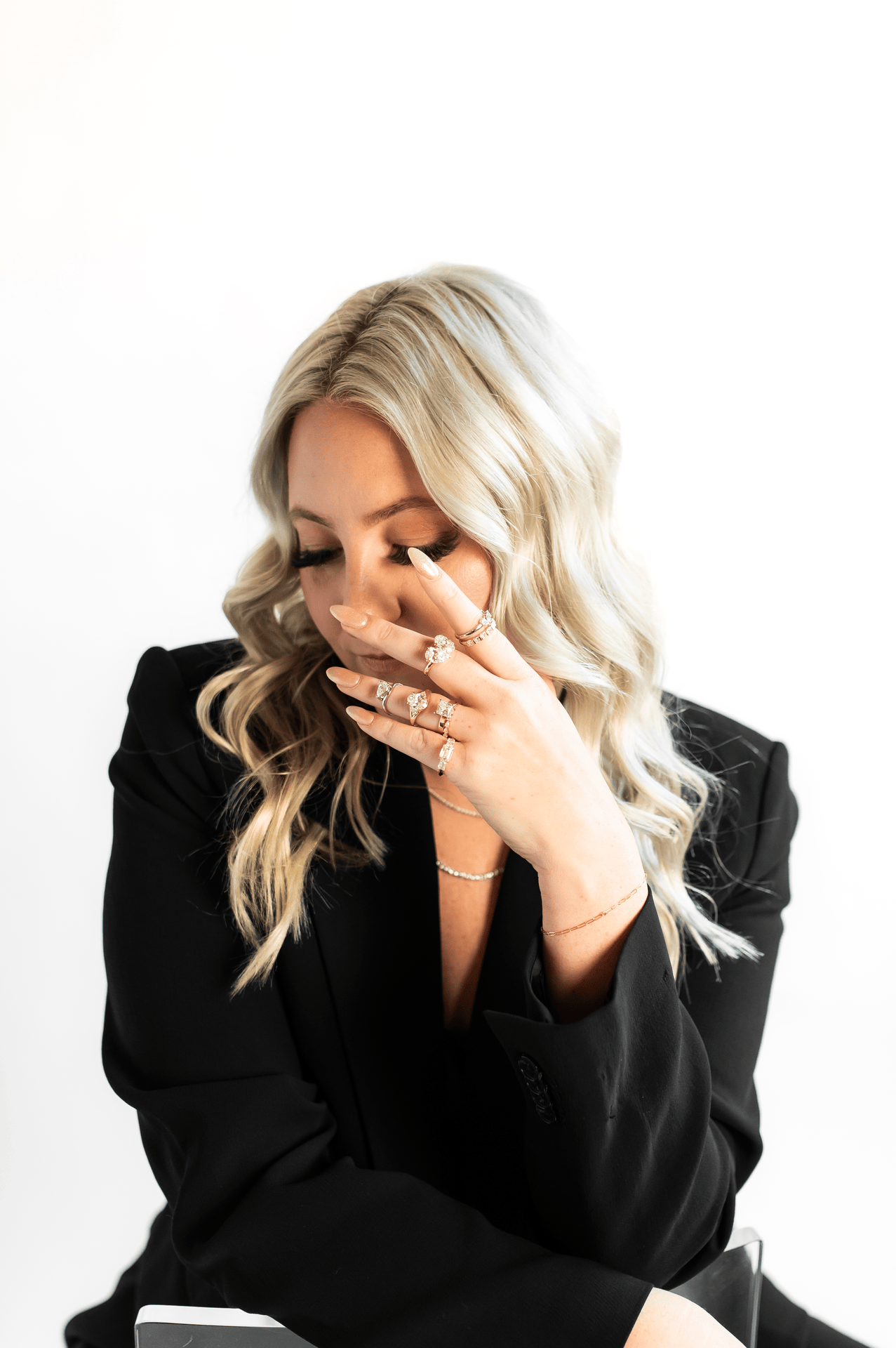A woman with long, wavy blonde hair wearing a black blazer and multiple rings on her fingers, touching her face with her eyes closed against a white background.