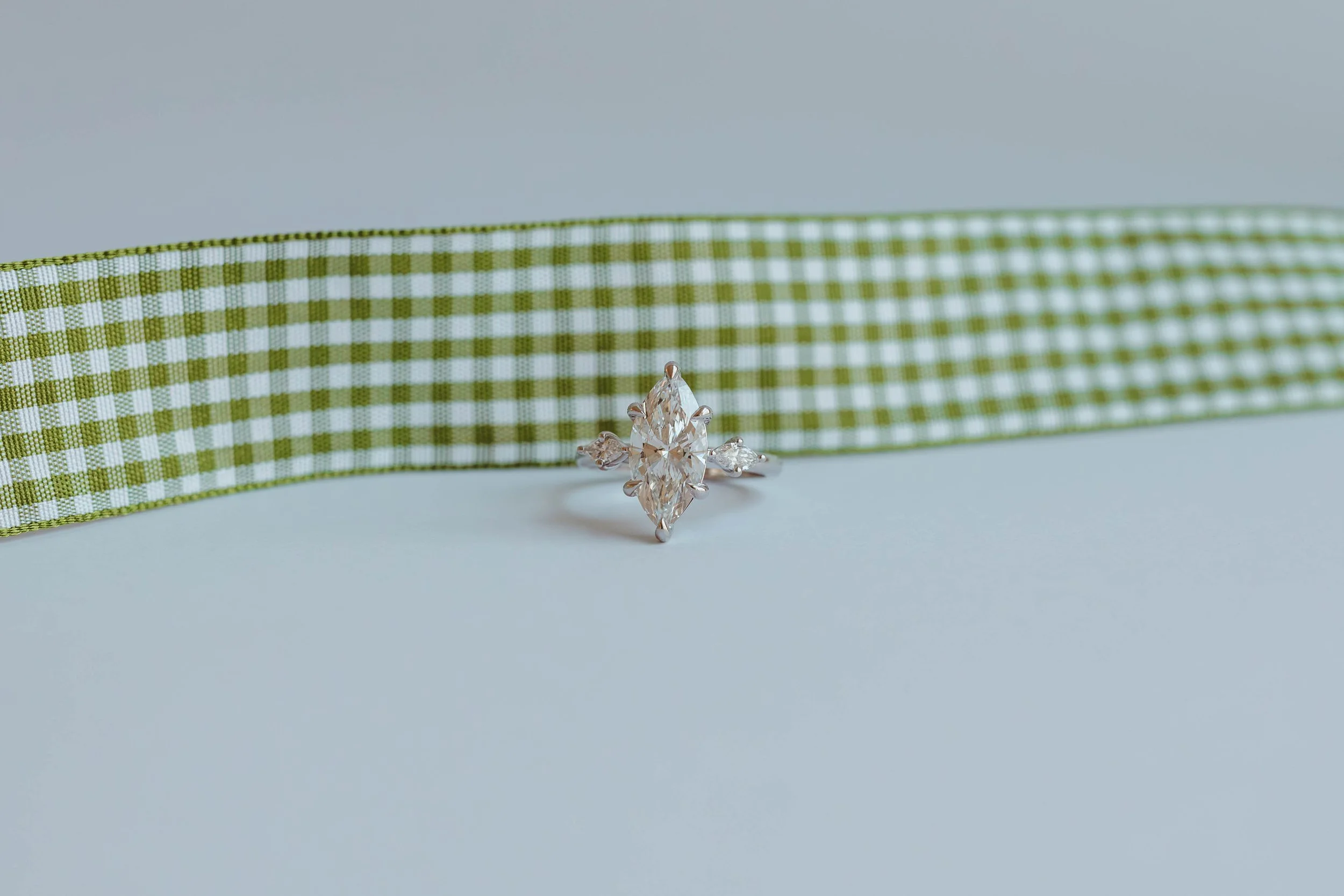 A silver ring with a marquise-cut central diamond and smaller diamonds on each side, resting on a light blue surface with a green and white gingham ribbon in the background.