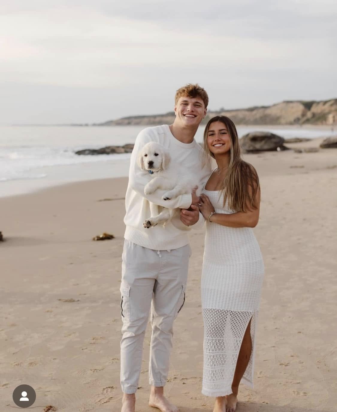 A smiling young man and woman stand on a sandy beach with an ocean and cliffs in the background. The man holds a fluffy white puppy, and they both wear light-colored casual clothes.