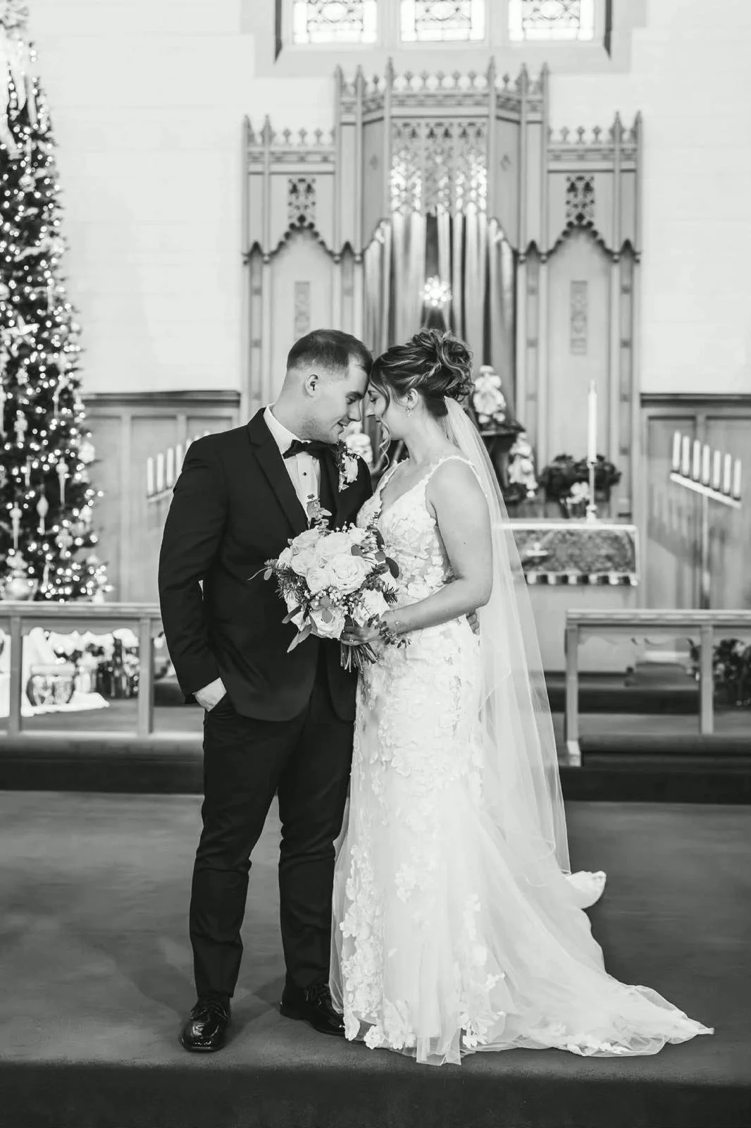 Black and white photograph of a newlywed couple standing close together inside a church, with their foreheads touching and eyes closed. The groom is dressed in a tuxedo and the bride in a lace wedding gown holding a bouquet of flowers.