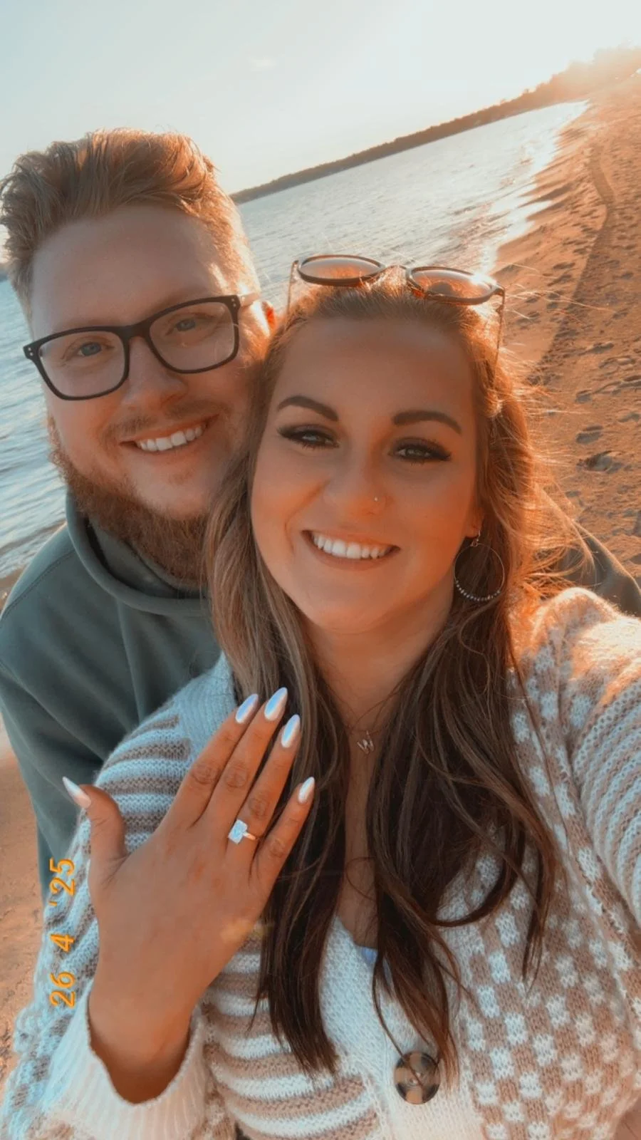A happy couple taking a selfie on a sandy beach near water during sunset, with the woman showing off her engagement ring.