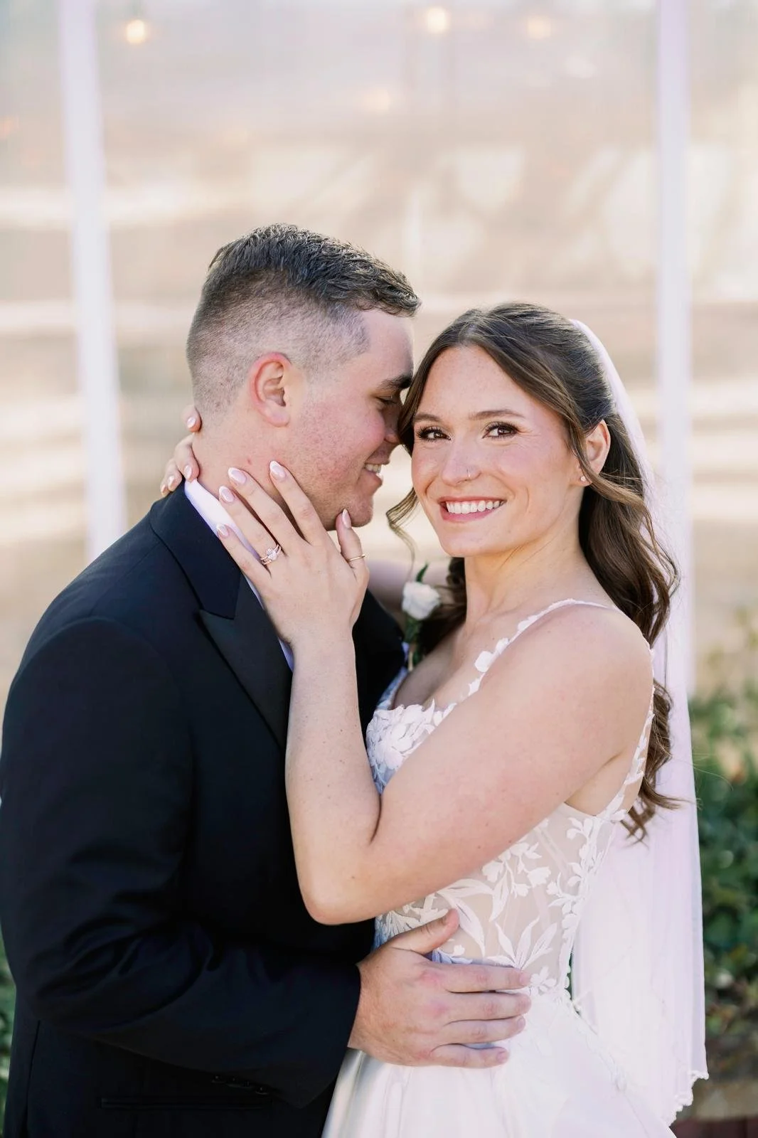 A newlywed couple smiling and embracing outdoors, with the woman's hand on the man's face, showing her engagement ring, and the man holding her waist.