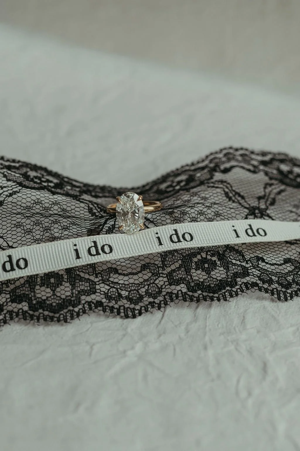 Diamond engagement ring resting on black lace fabric, with an 'i do' ribbon across it.