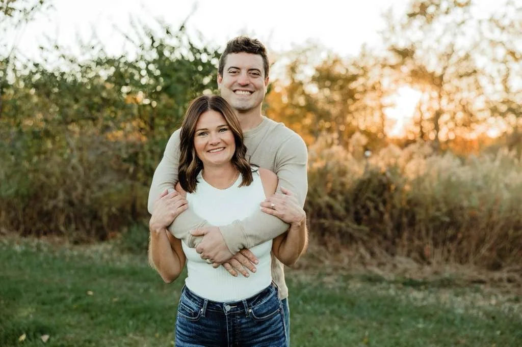 A smiling couple standing outdoors at sunset, with trees and foliage in the background.