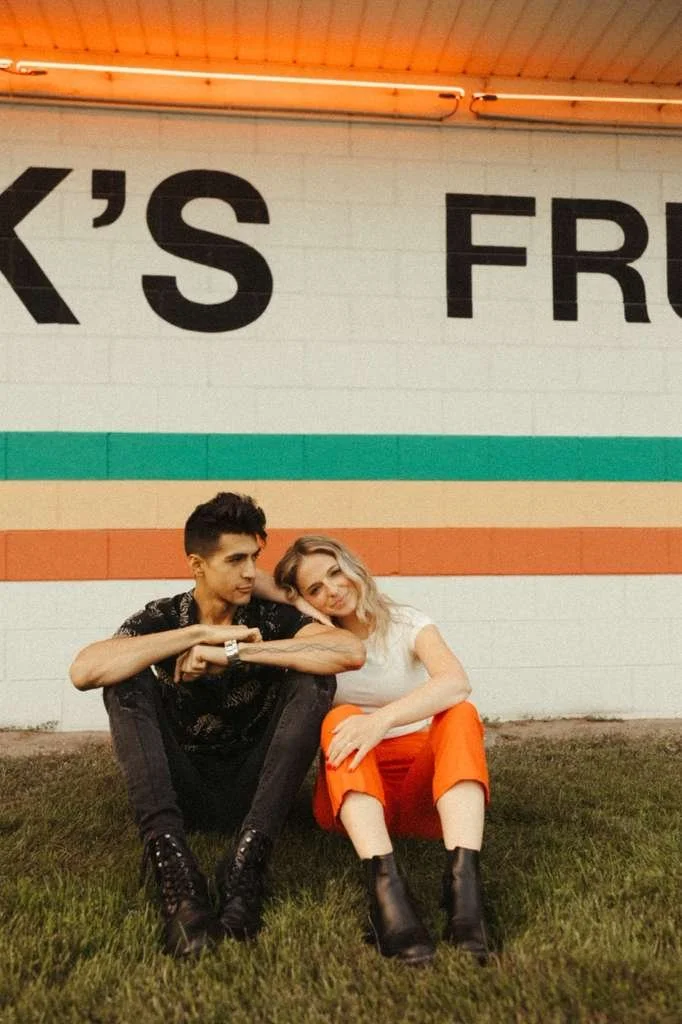 A young man and woman sitting on the grass in front of a colorful mural with the partial text 'K'S FRU' visible. They are leaning on each other, with the woman resting her head on the man's shoulder.