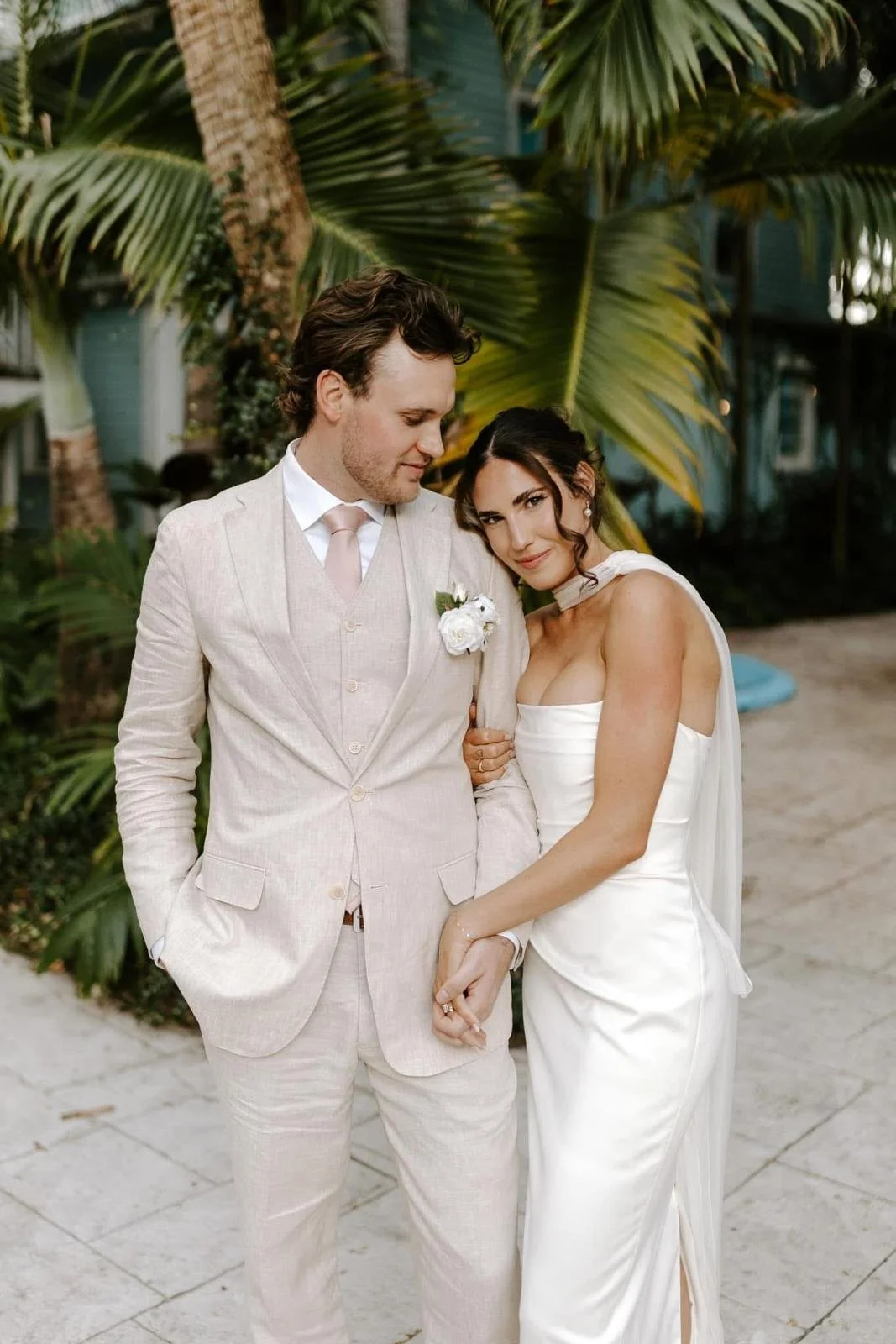 A bride and groom standing close together outdoors, surrounded by palm trees, during their wedding. The groom is wearing a light-colored suit with a boutonniere, and the bride is in a white wedding dress, holding hands and leaning her head on his shoulder.