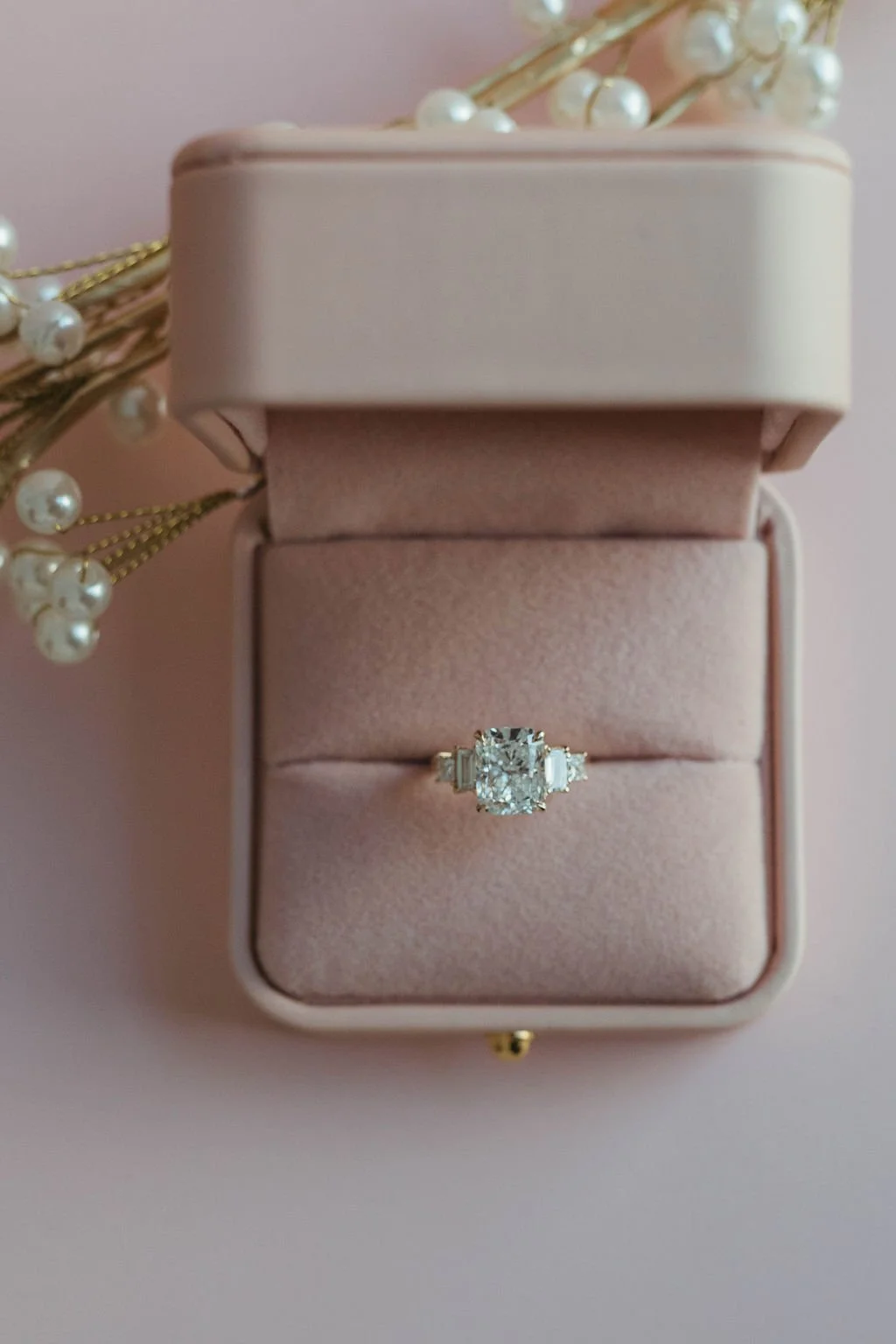 A diamond engagement ring in a pink velvet ring box, with a pearl and gold embroidery decoration beside it.