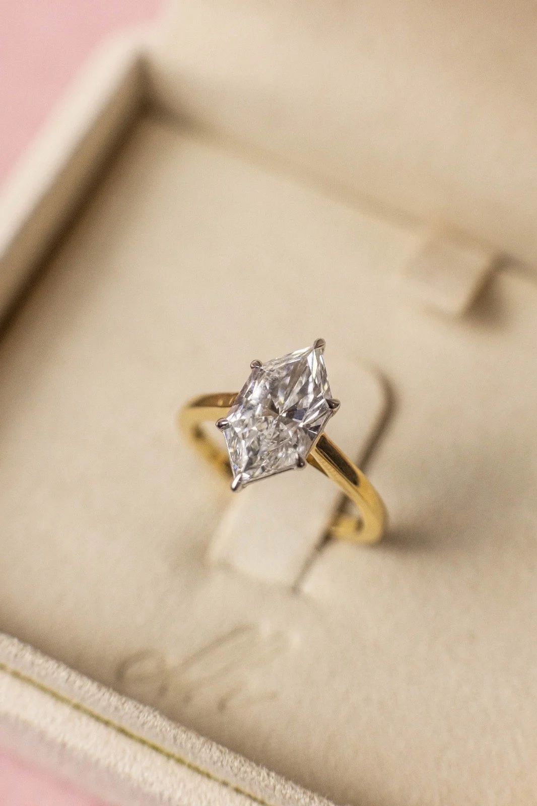 A gold ring with a large princess-cut diamond, resting inside a jewelry box with a pink exterior and a beige interior.