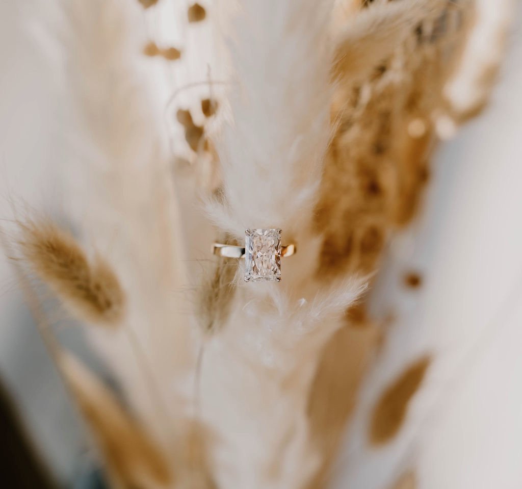 Close-up photo of a ring with a rectangular diamond, set in gold, placed against a soft, textured background with beige and brown tones.