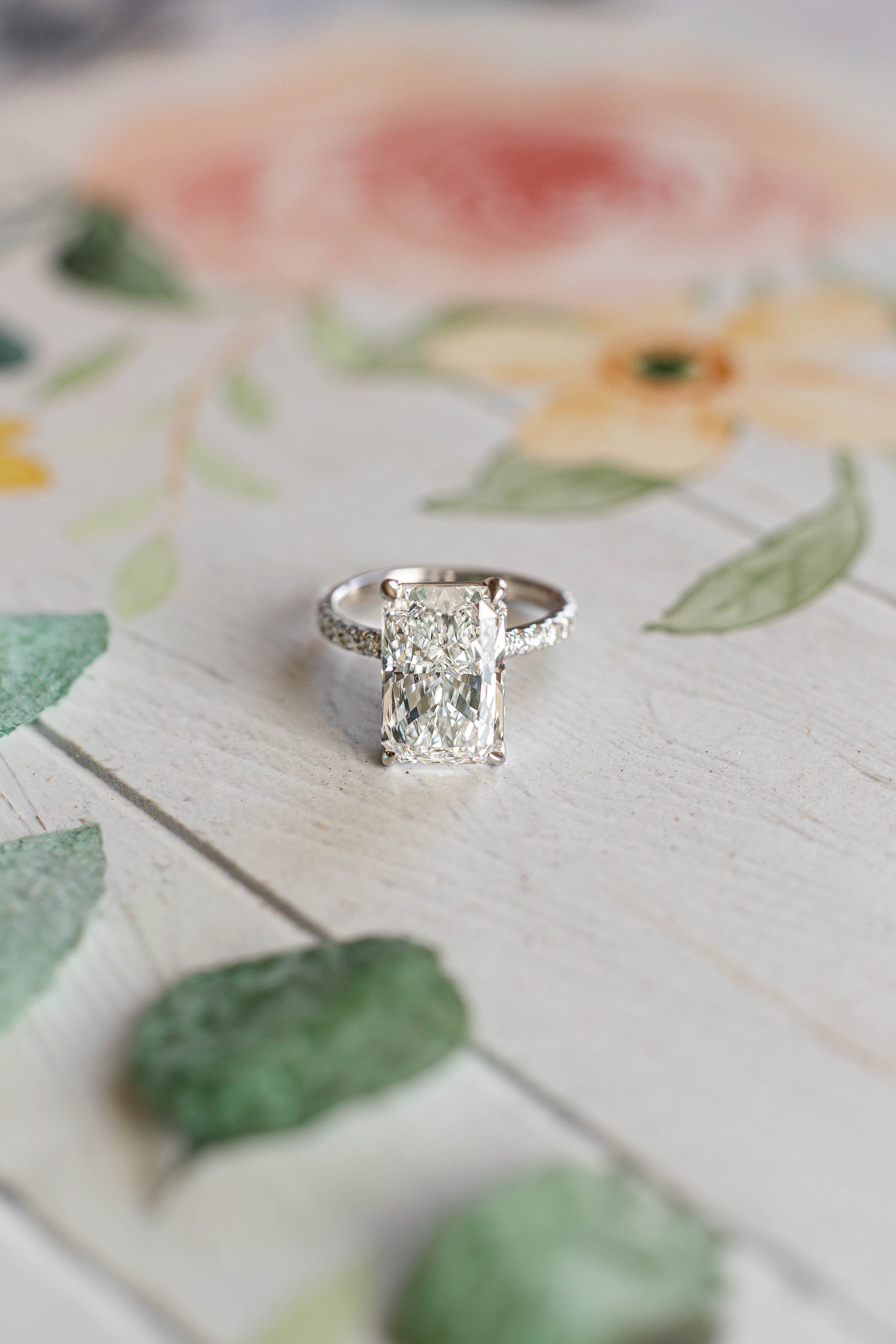 A silver ring with a large rectangular clear gemstone, possibly a diamond, resting on a light-colored wooden surface decorated with floral and leaf patterns.