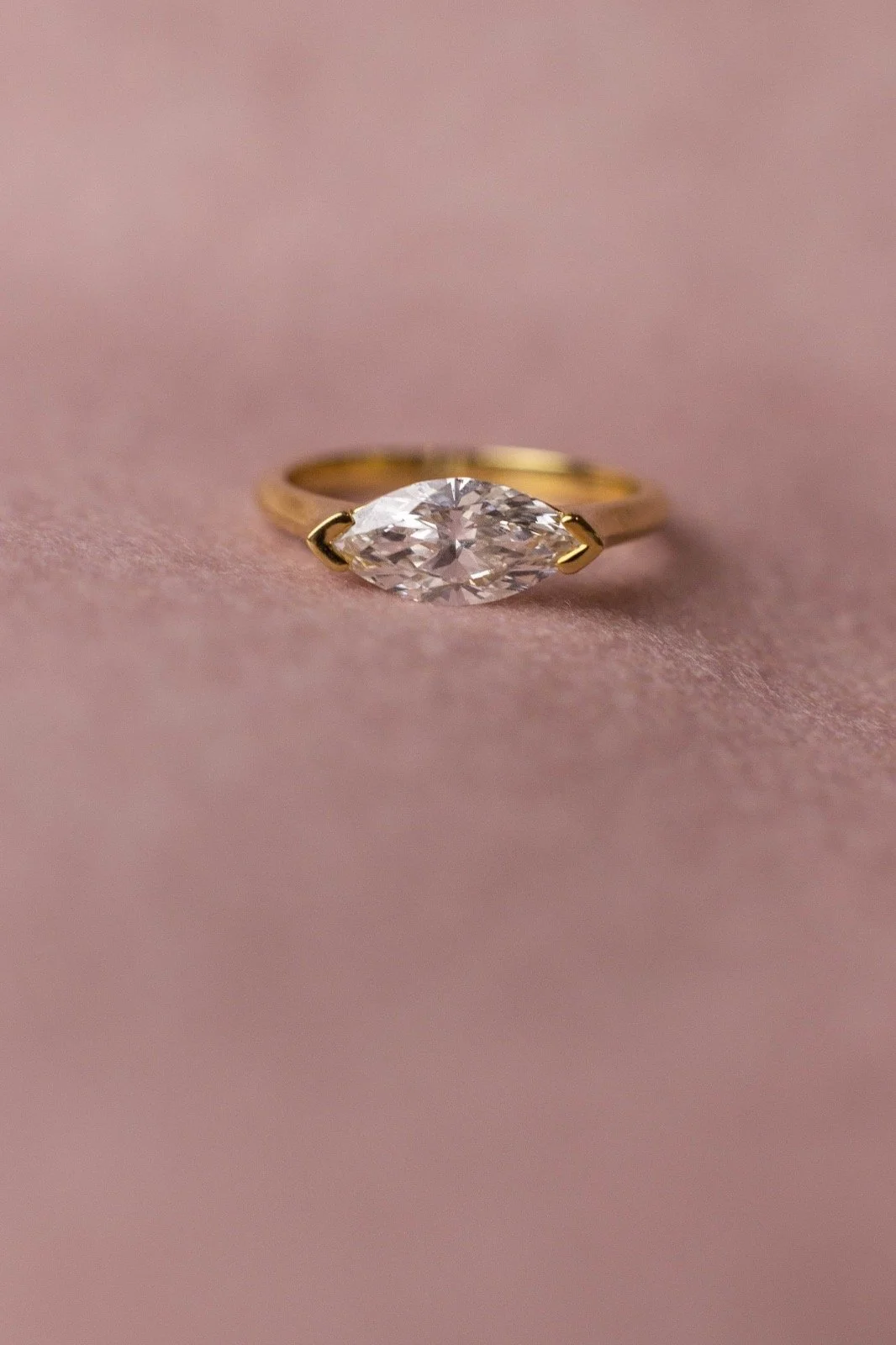 A gold ring with a marquise-cut diamond, resting on a pink fabric surface.