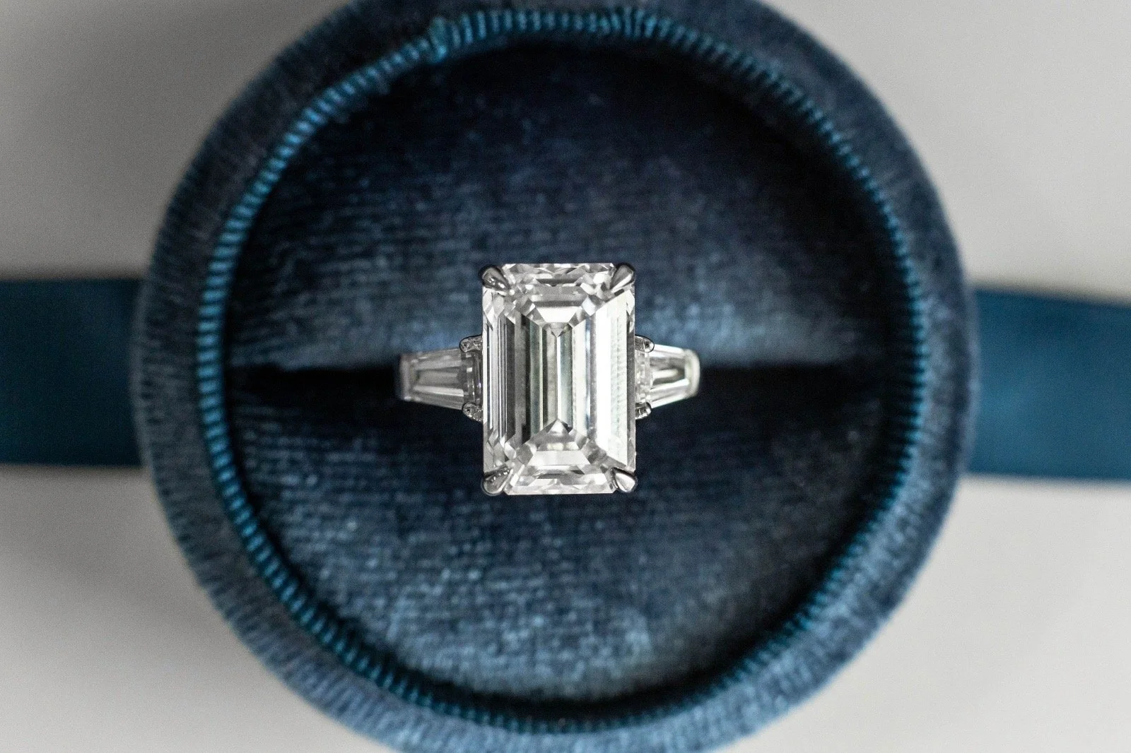 A silver ring with a large rectangular emerald-cut diamond in a black jewelry box with a dark interior and a light gray background.