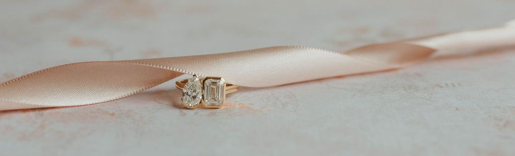 Close-up of a pink satin ribbon tied around two sparkling engagement rings on a soft surface.