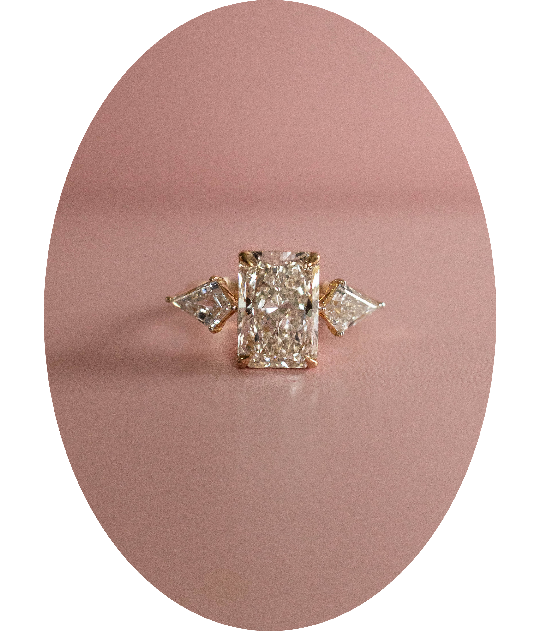 Close-up of a diamond ring with a rectangular center stone and two triangular side stones on a pink surface.