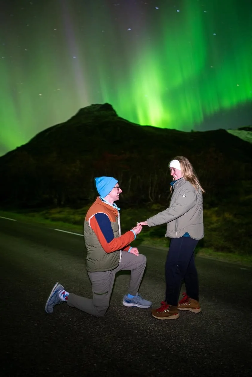 A man is proposing to a woman on a road at night with the Northern Lights illuminating the sky behind them, mountain silhouette in the background.