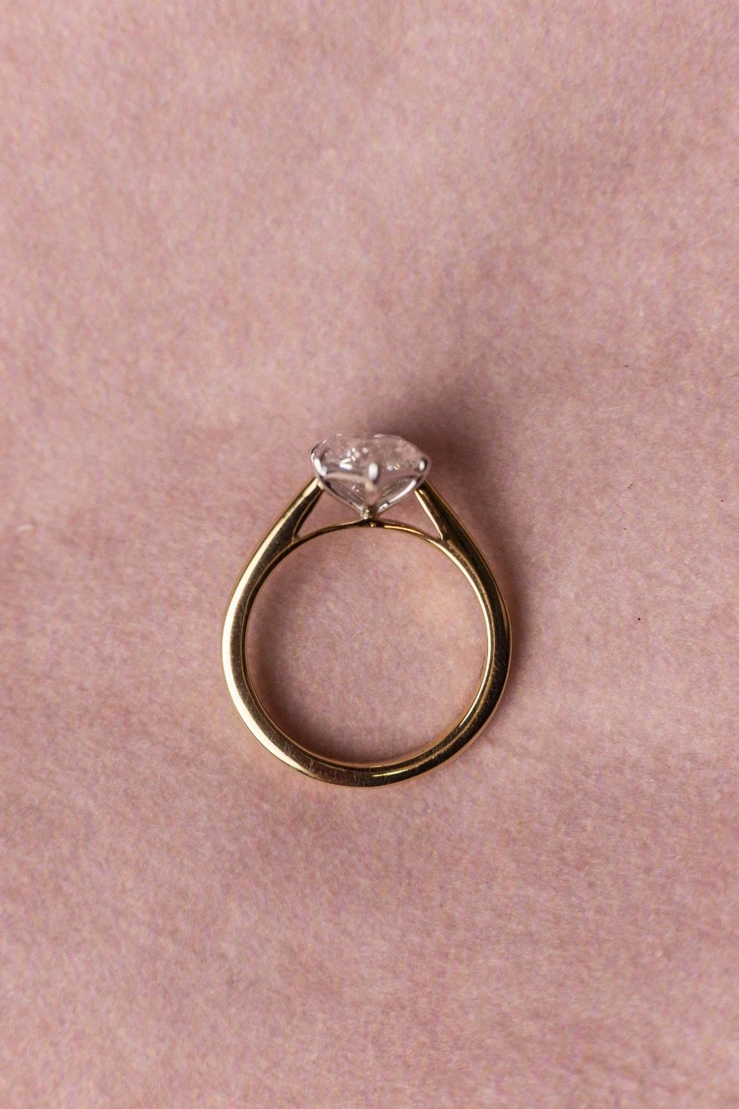 A gold engagement ring with a large, sparkling diamond solitaire, placed on a soft pink textured background.