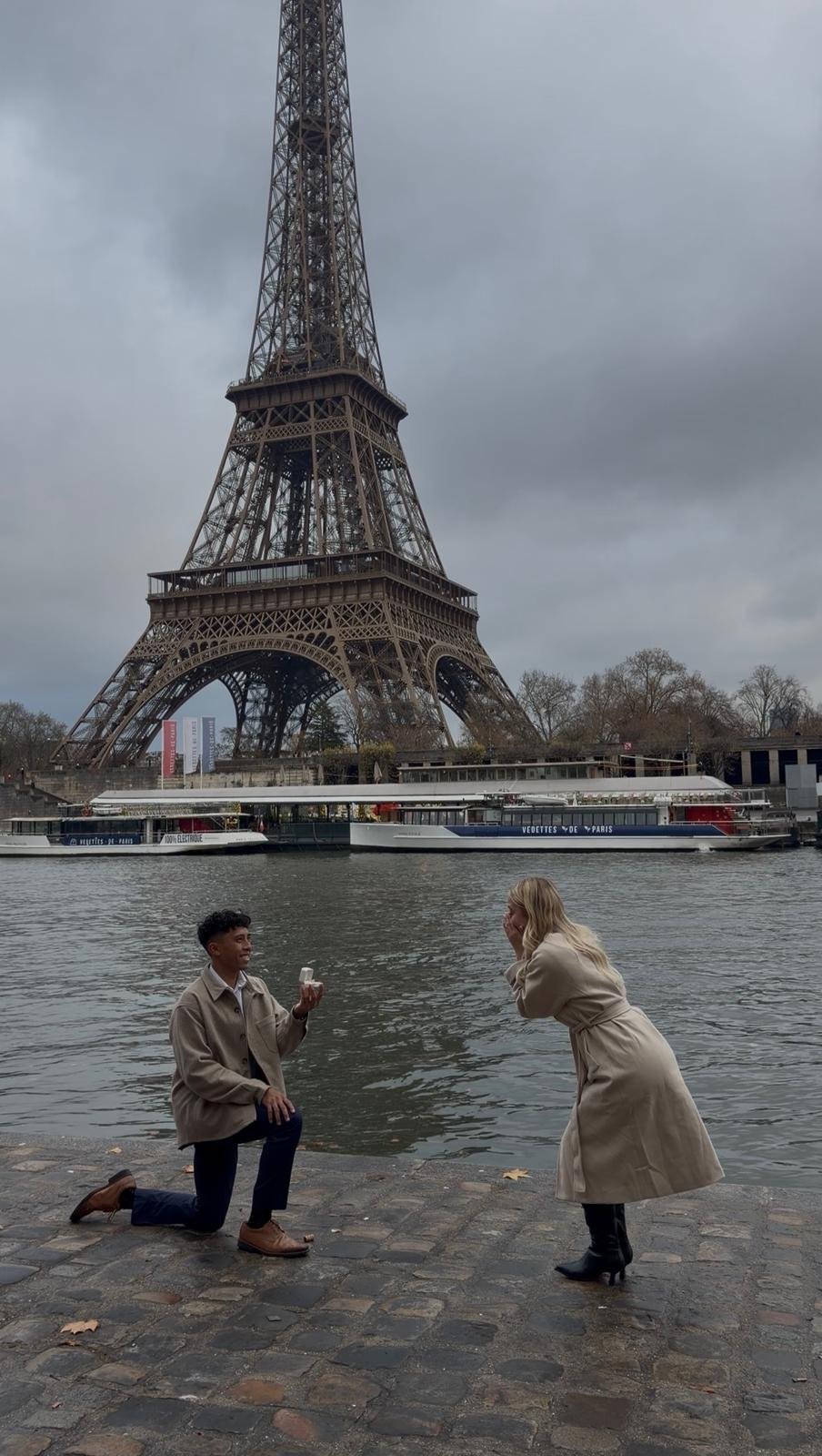 A man on one knee proposing to a woman by the water with the Eiffel Tower in the background on a cloudy day.