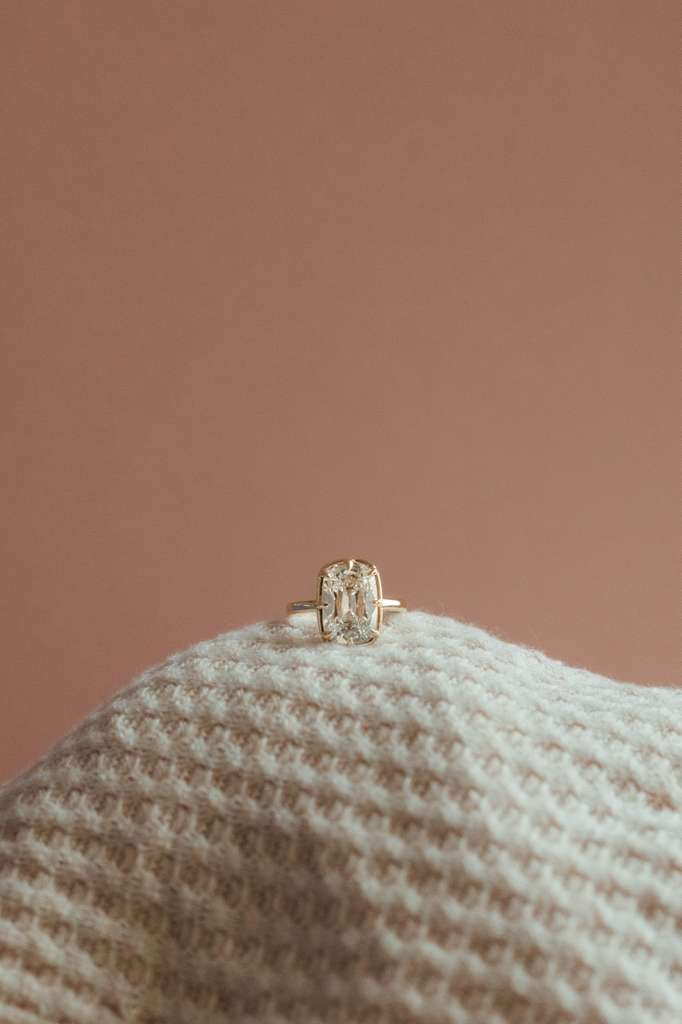 A close-up of an elegant diamond ring on a soft, textured beige surface against a plain pink background.